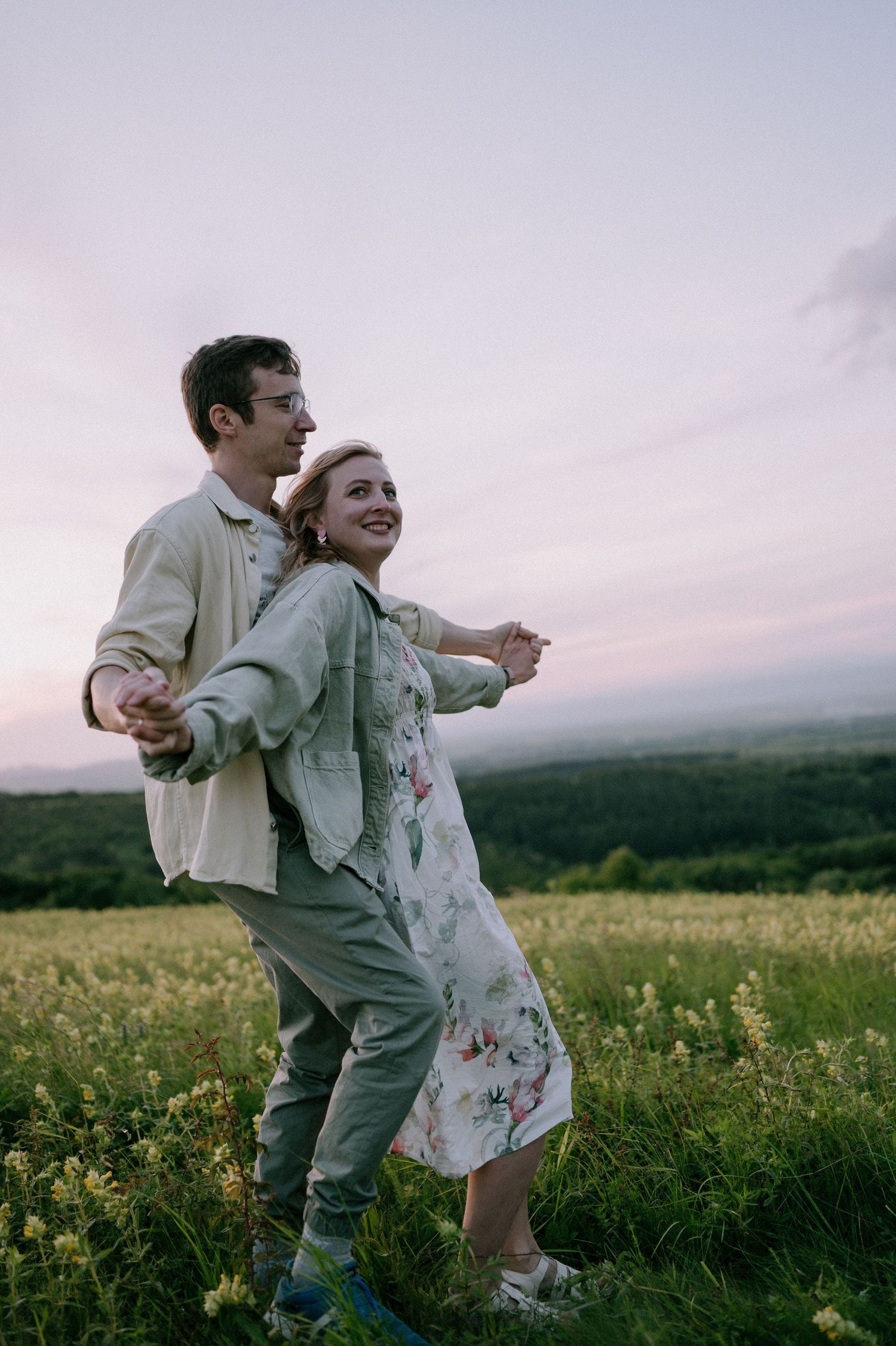 Two people dance and laugh together in a flowering field with mountains in the distance.