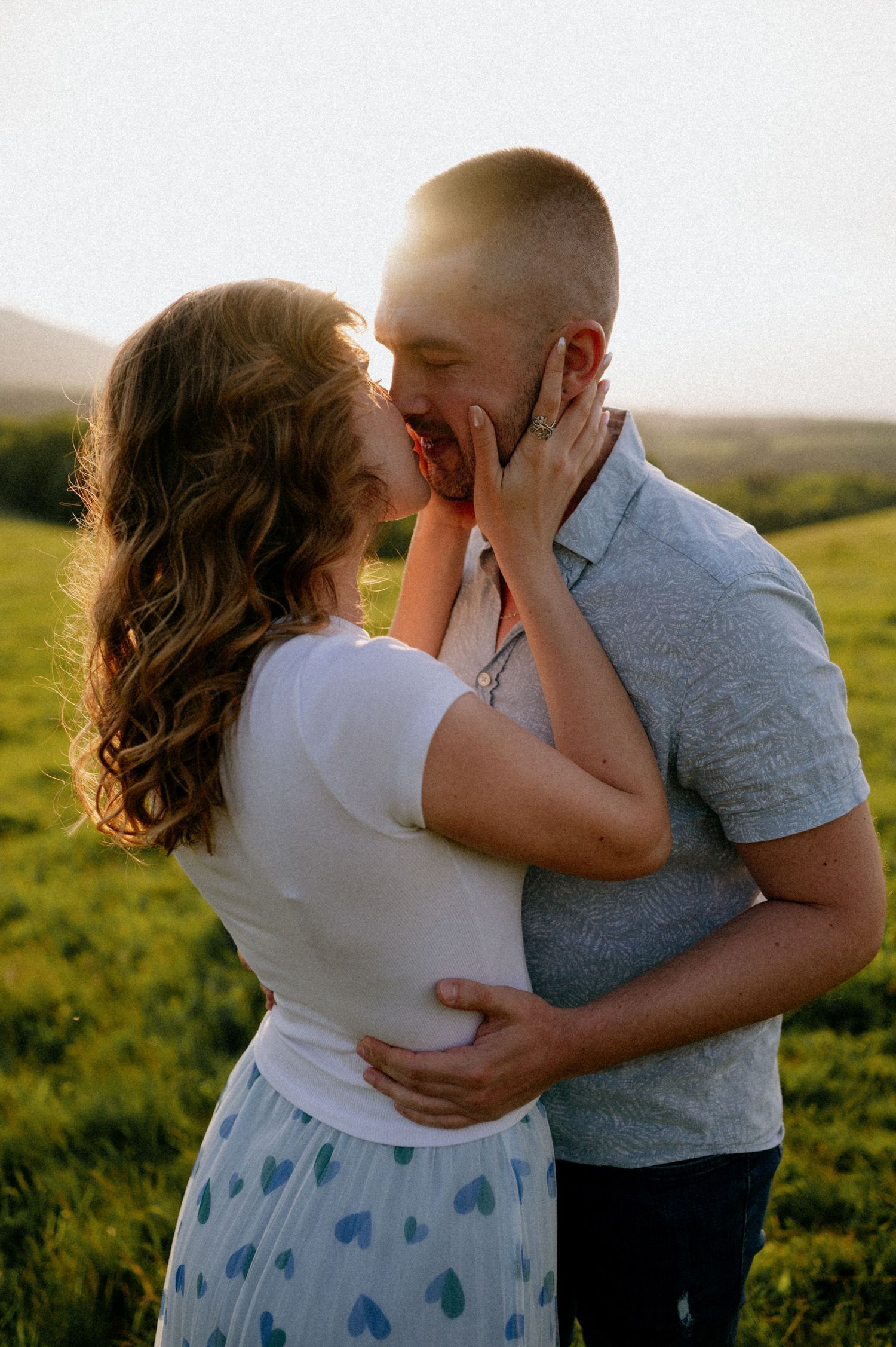 A couple shares an intimate embrace against a backdrop of rolling hills at sunset.