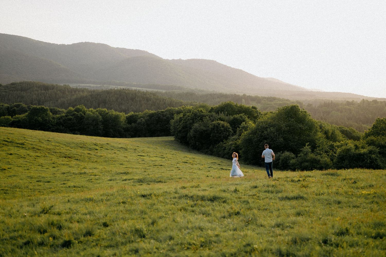 Two figures walk across a sunlit grassy hillside with mountains and woodland in the background.