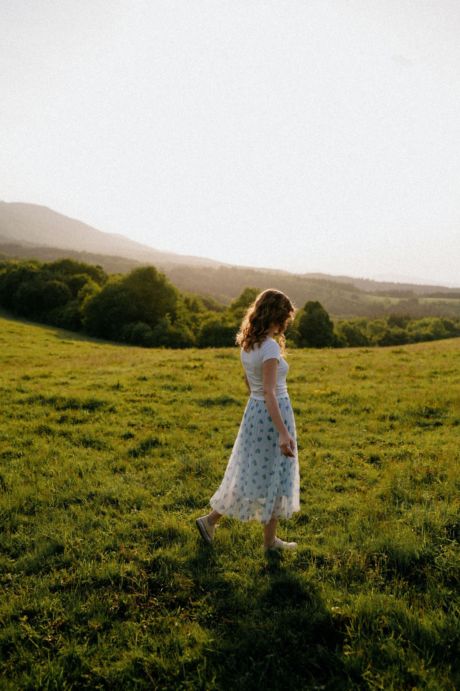 A figure in a flowy white dress twirls gracefully across a sunlit hilltop meadow at golden hour.