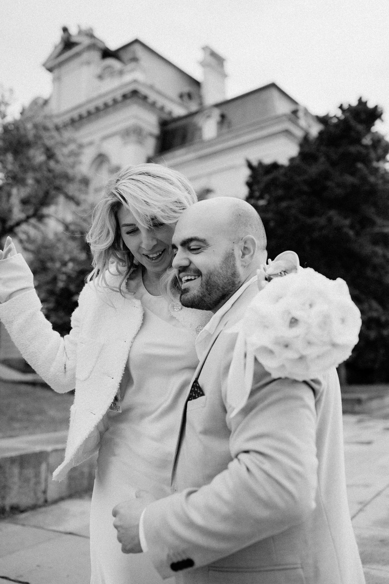 Two people in white outfits laugh and embrace joyfully near a historic building.