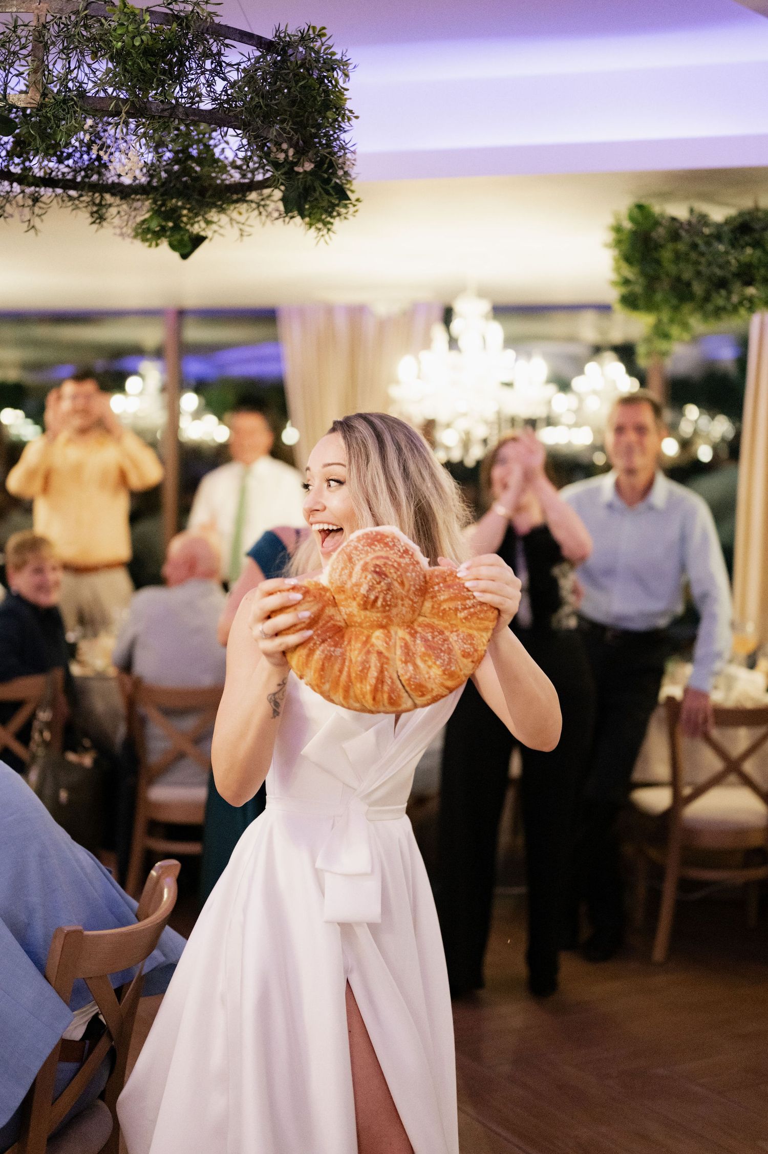 A person in a white wedding dress holds up a large croissant while celebrating at an elegant reception venue.