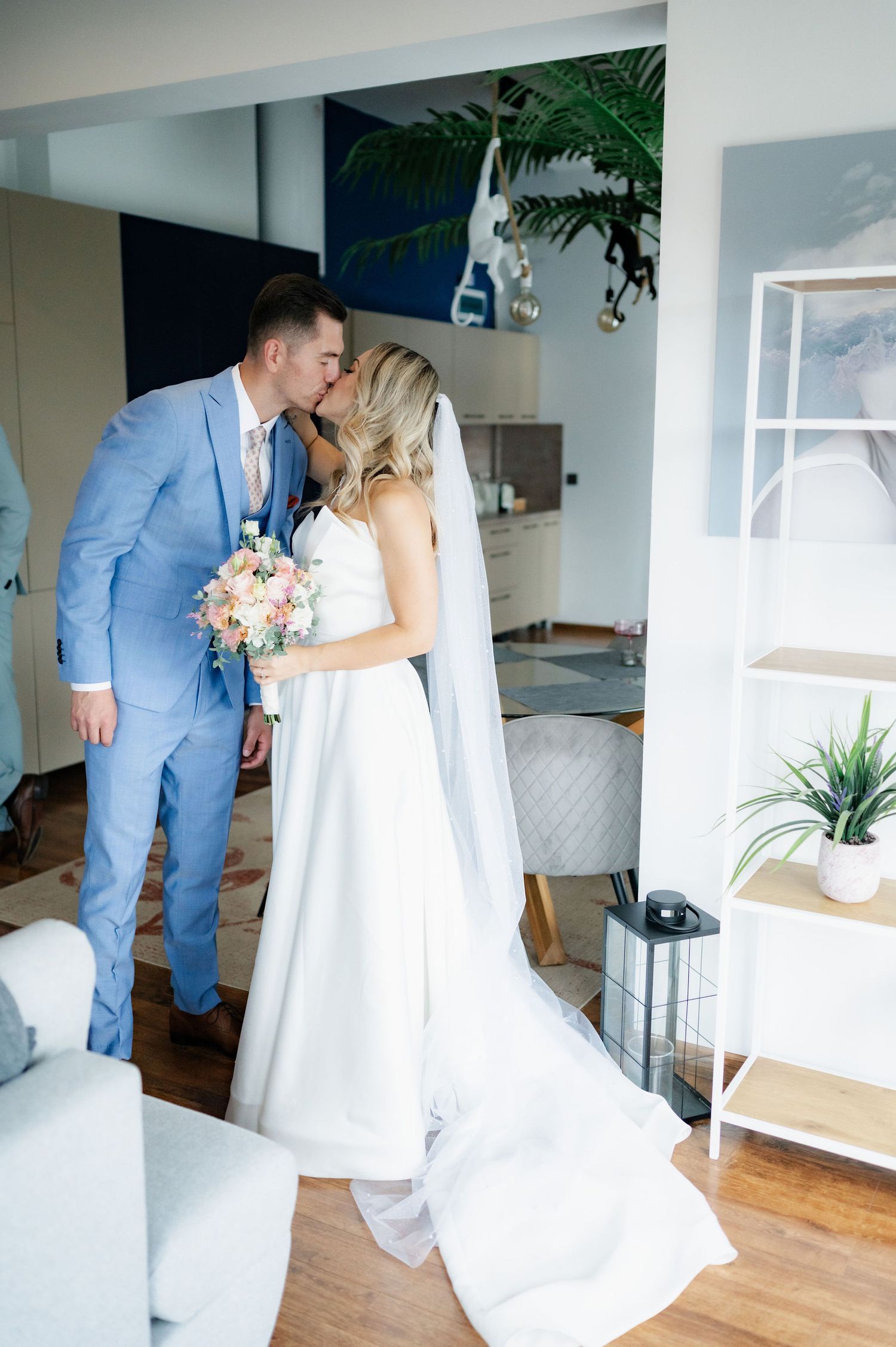 A couple in wedding attire share intimate moments in a bright indoor setting with white walls and wooden floors.