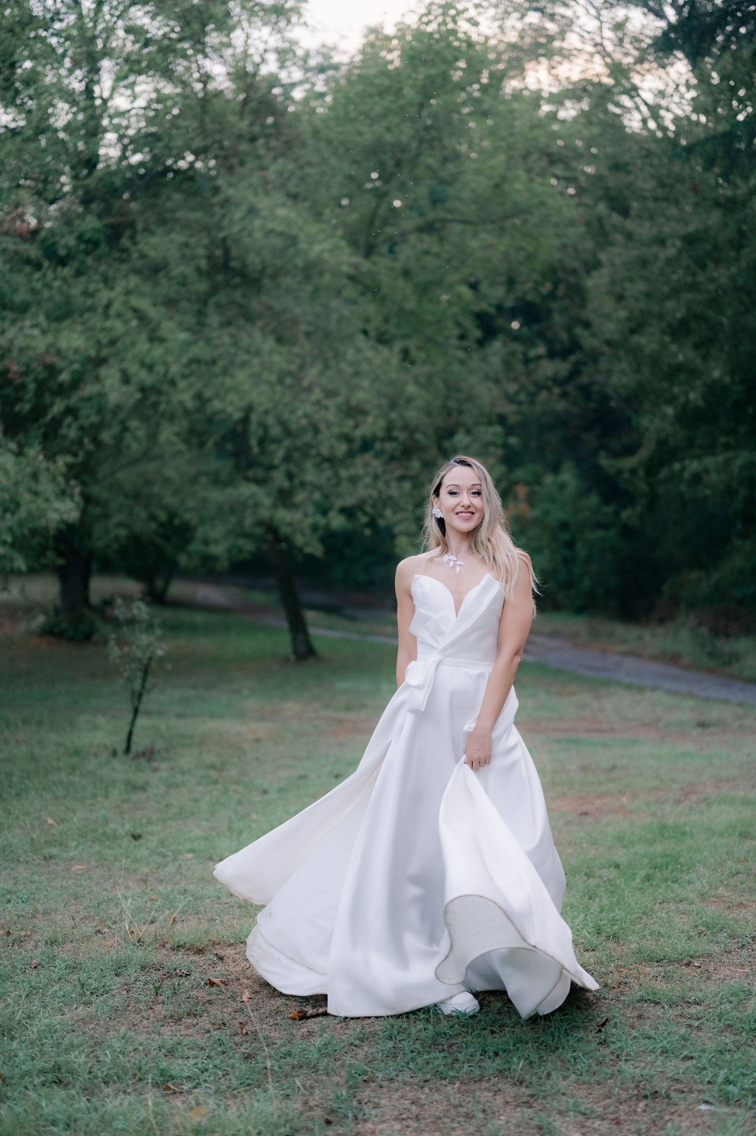 A person in a flowing white wedding dress twirls and dances playfully in a green outdoor park setting at dusk.
