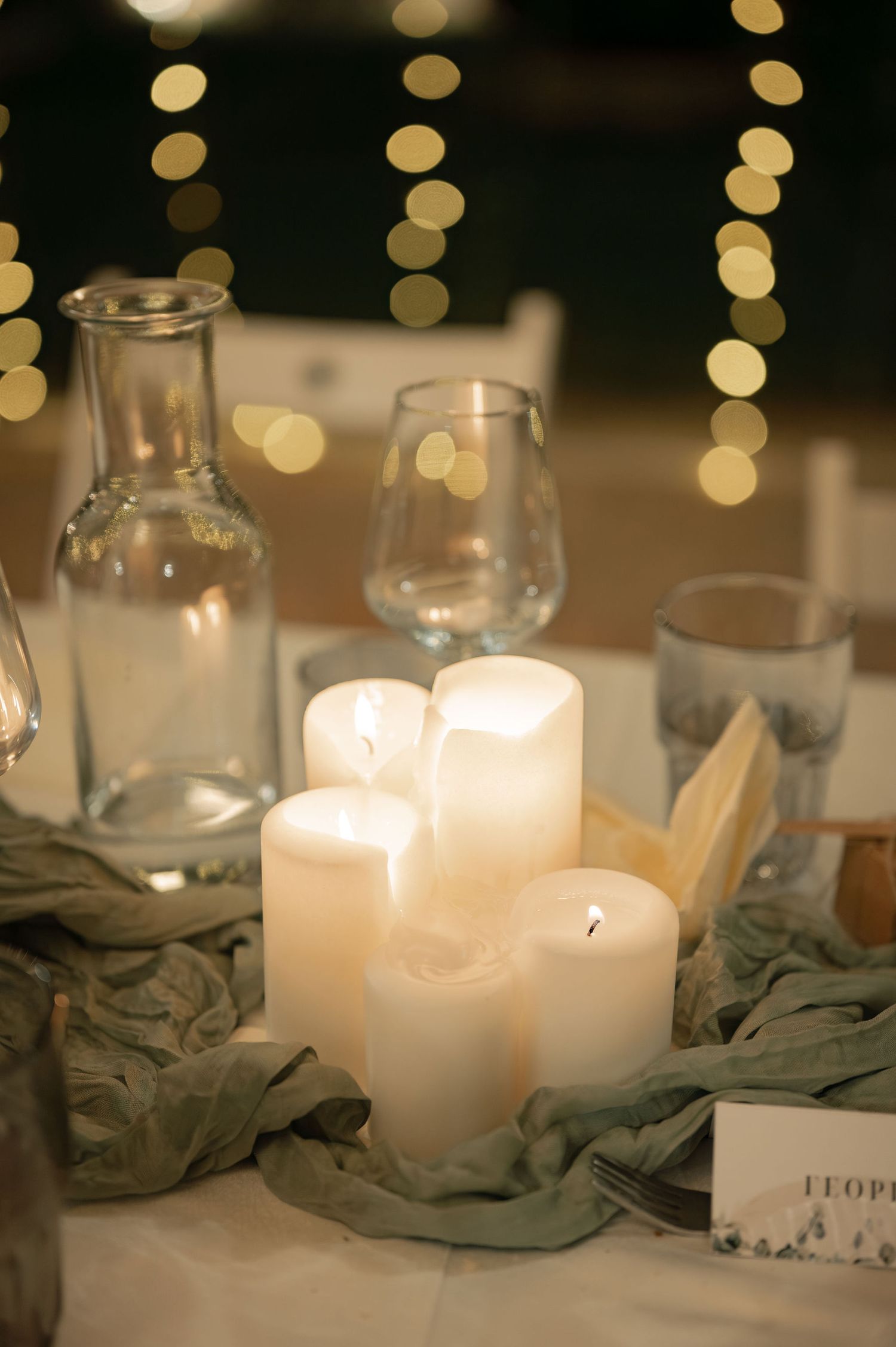 White pillar candles and soft fabric create an elegant table setting with bokeh lights in the background.