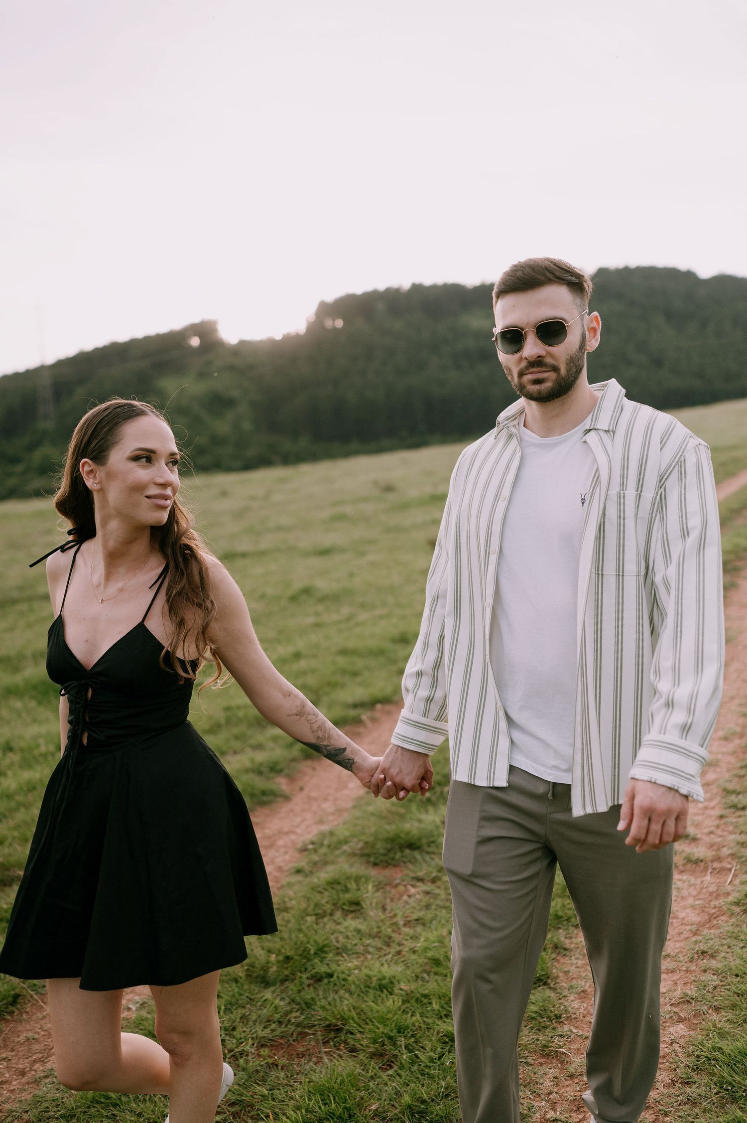 A couple in casual attire shares playful moments while walking through a grassy hillside at sunset.