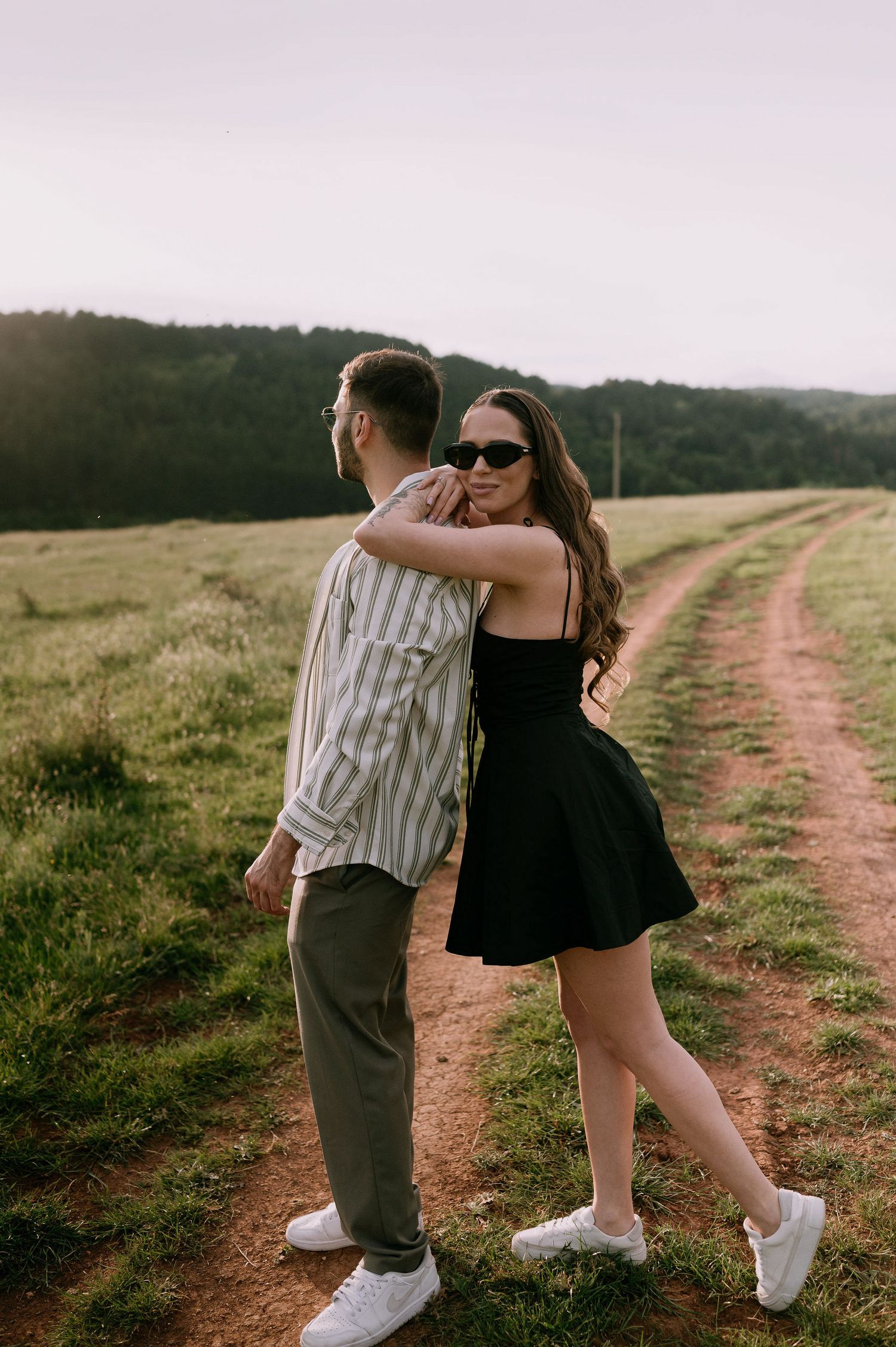 A couple embraces on a dirt path through rolling hills during golden hour, with one wearing a black dress and another in stripes.