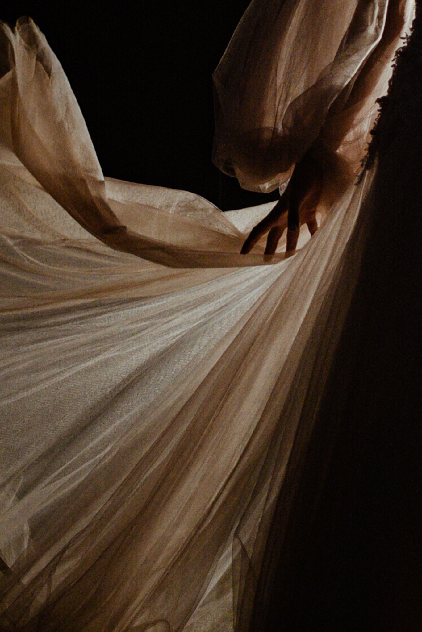 Hands delicately hold and spread flowing white tulle fabric in an artistic arrangement against a dark background.