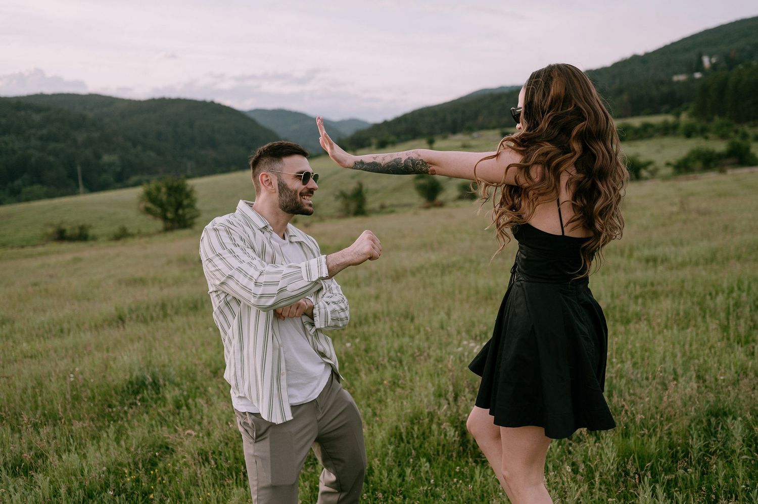 A couple dances playfully in a grassy meadow with rolling green hills and mountains in the background at sunset.