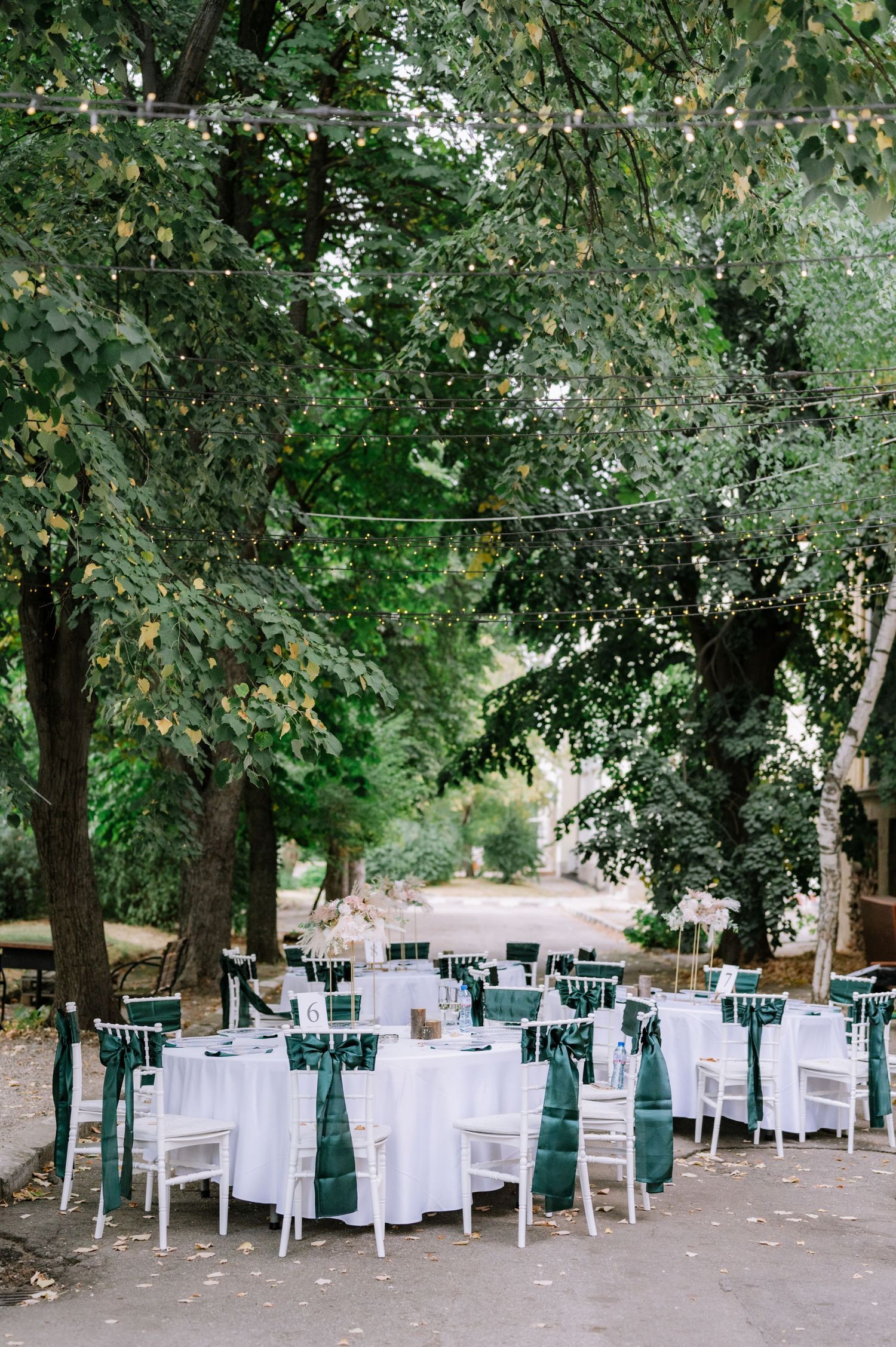 White tables and chairs arranged for outdoor dining under shady trees in a tranquil garden setting.