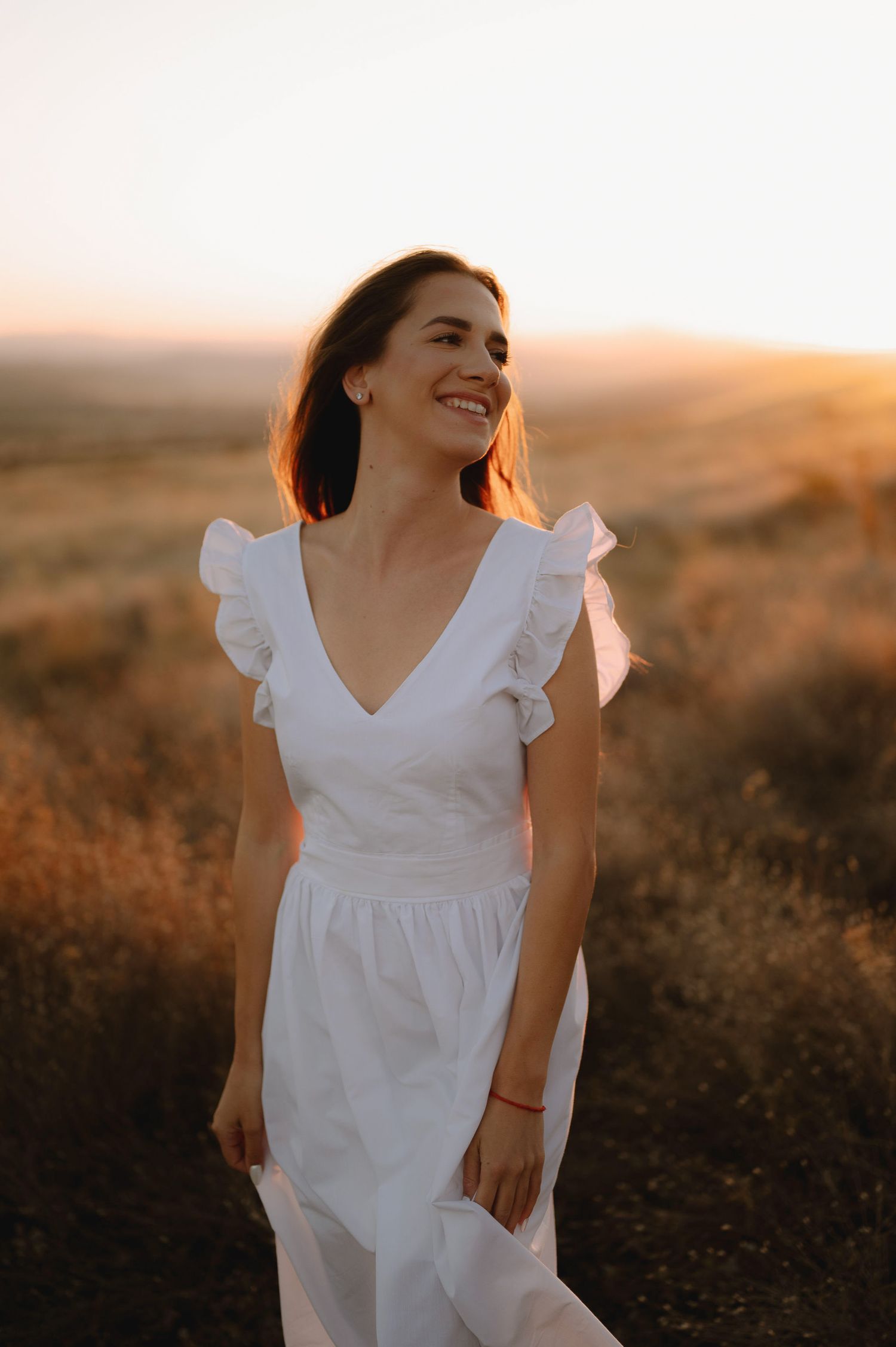 Person in flowing white dress poses joyfully in golden sunlit field during sunset.