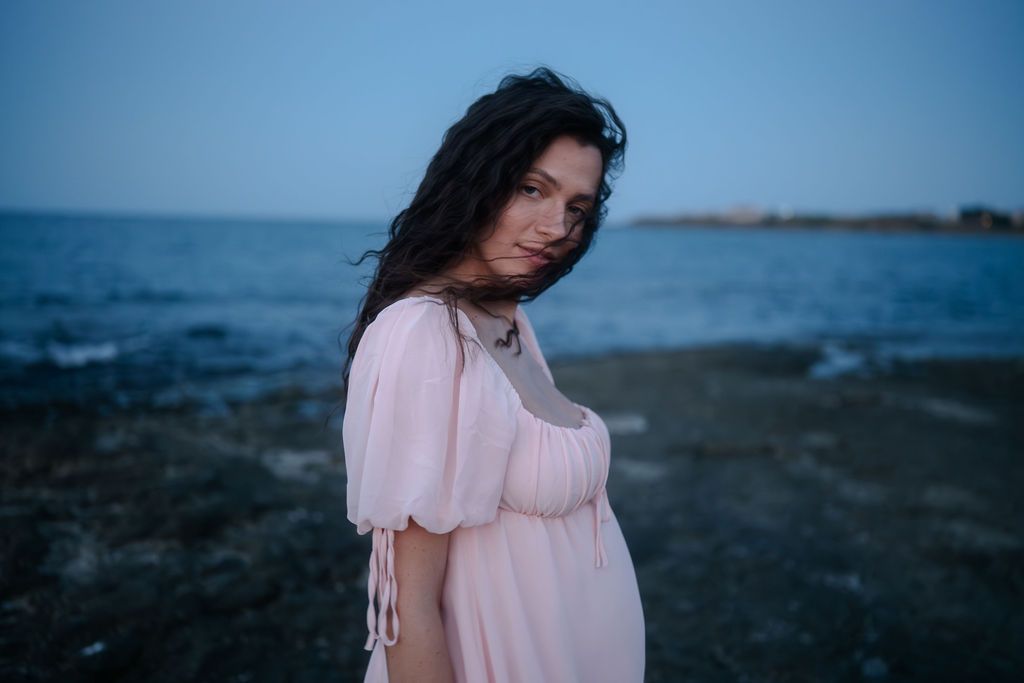 A person in a pink dress stands thoughtfully by the ocean shore at dusk against a moody blue sky.