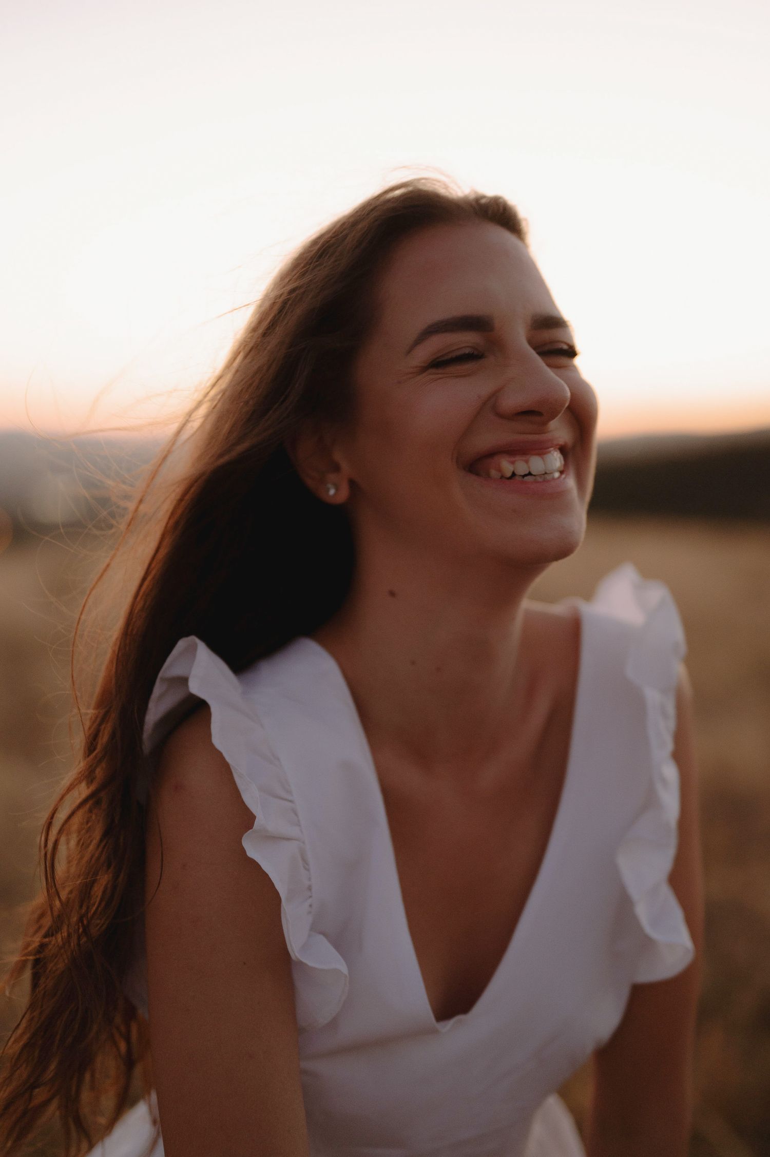 A woman in a white ruffled top smiles joyfully while standing outdoors during golden hour sunset.