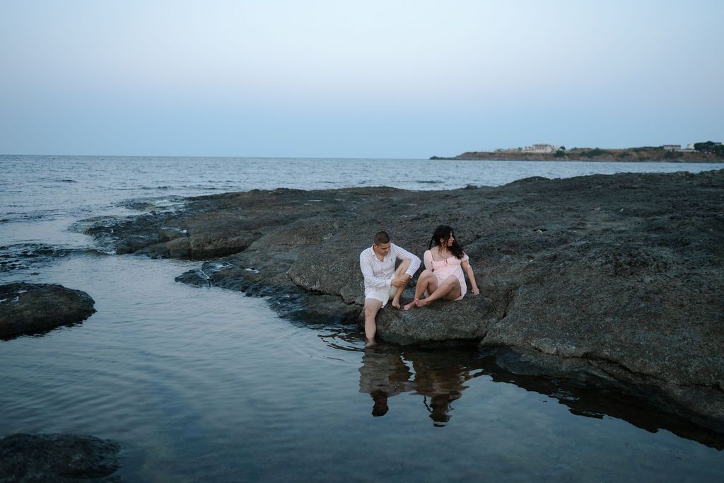 Couple sitting together on rocky coastal shoreline during sunset creating reflections in the water.