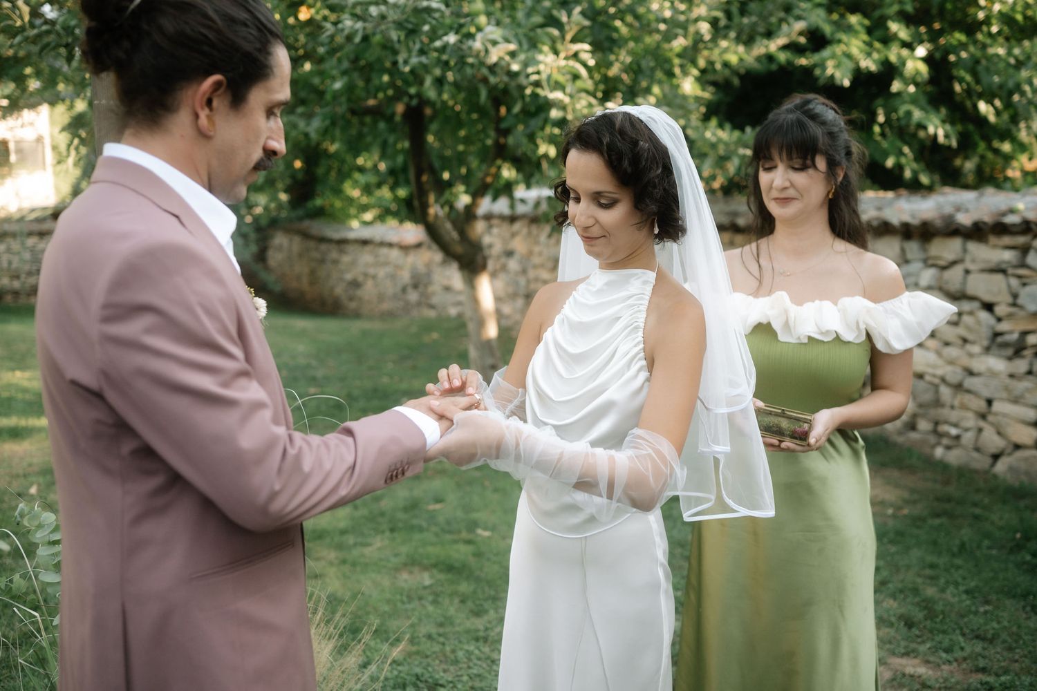 A wedding ceremony takes place in a garden setting with the couple exchanging rings as a bridesmaid in sage green looks on.
