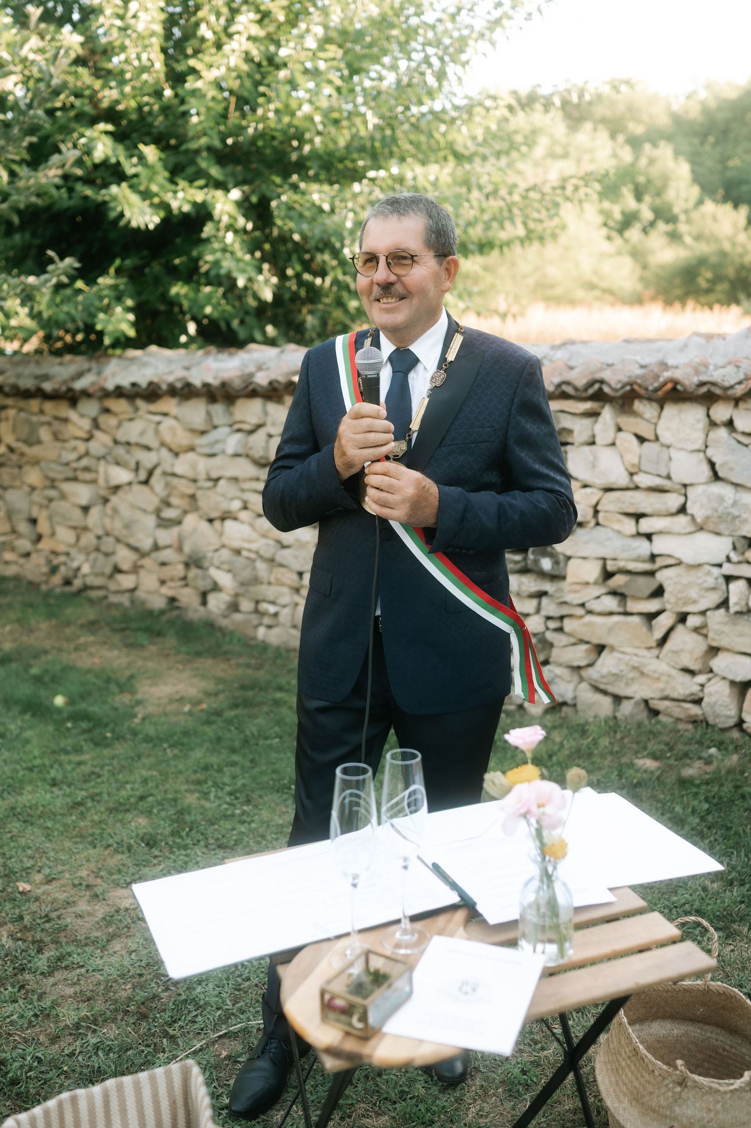 Official in dark suit stands at outdoor stone wall signing documents at rustic table with glass of champagne.