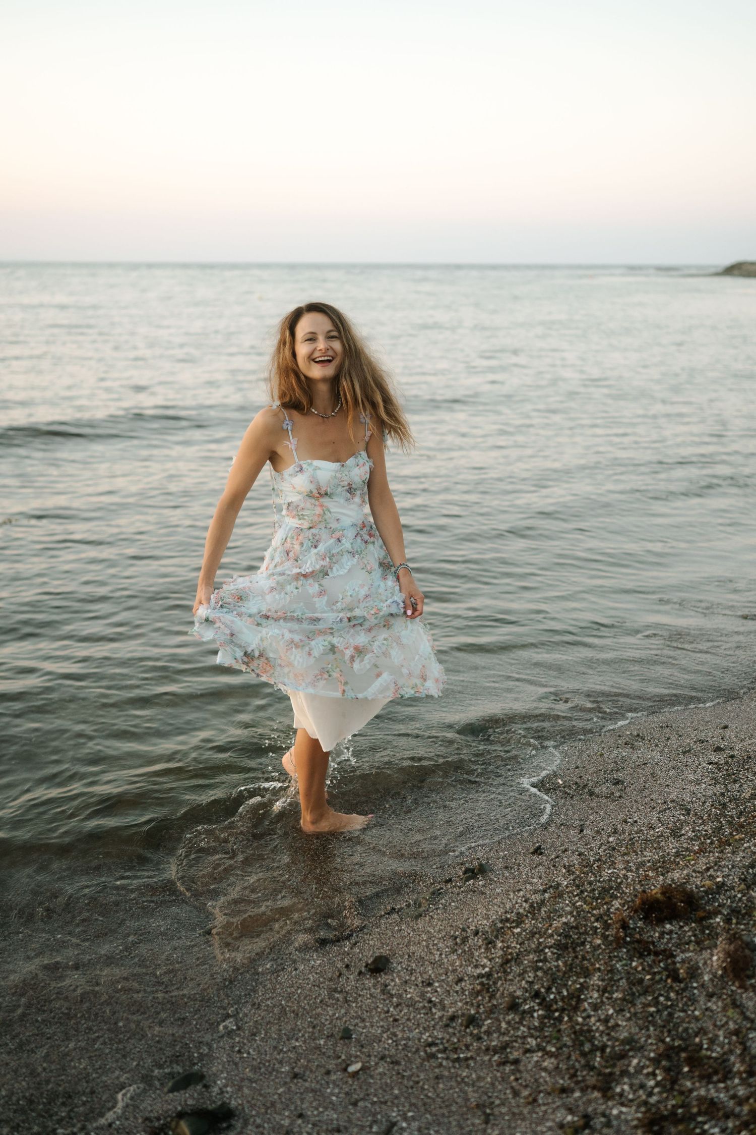A person in a flowing floral dress splashes and plays along the rocky shoreline at sunset.