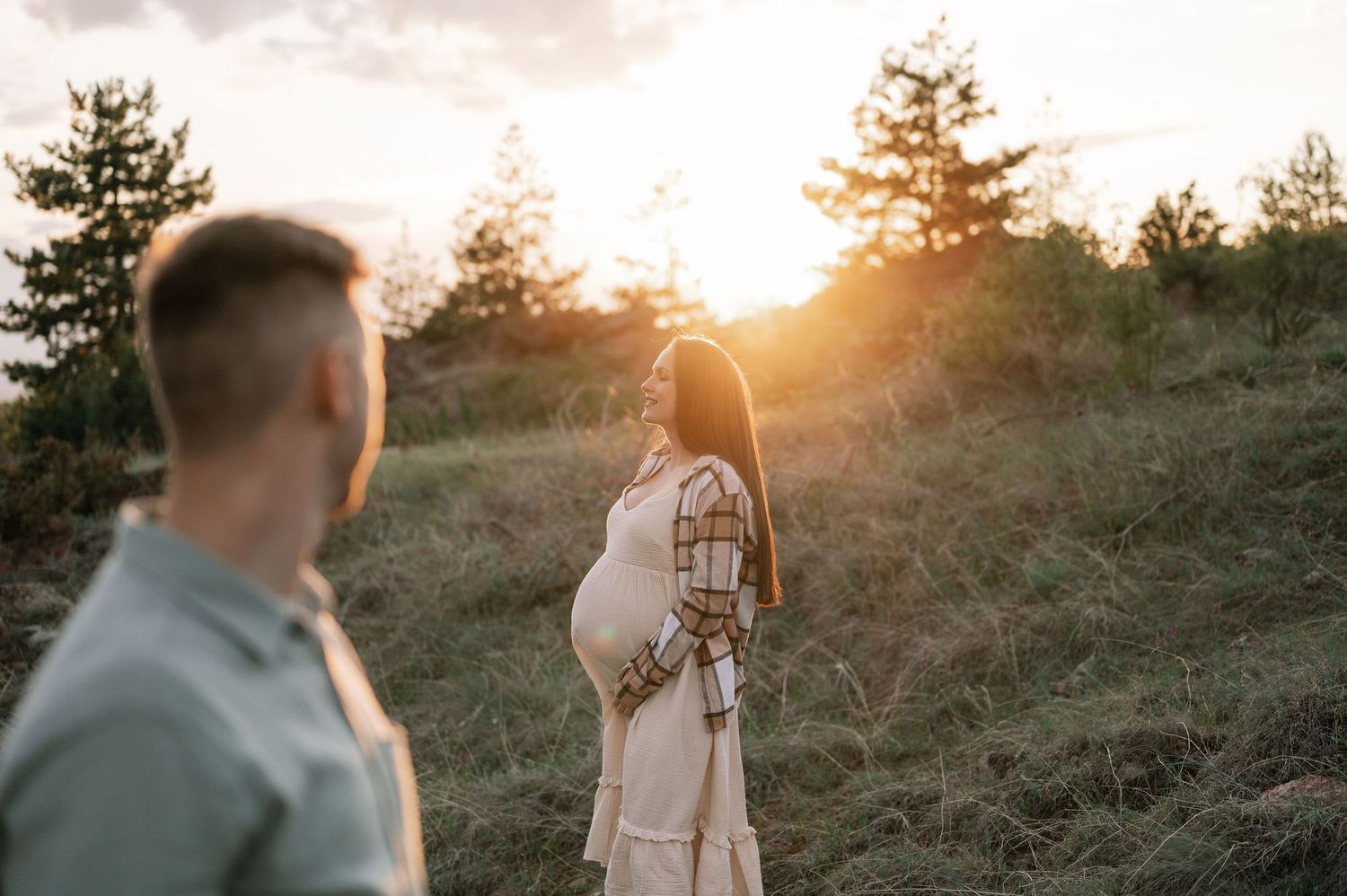 A couple shares a romantic moment in a sunlit meadow during golden hour while pine trees frame the background.