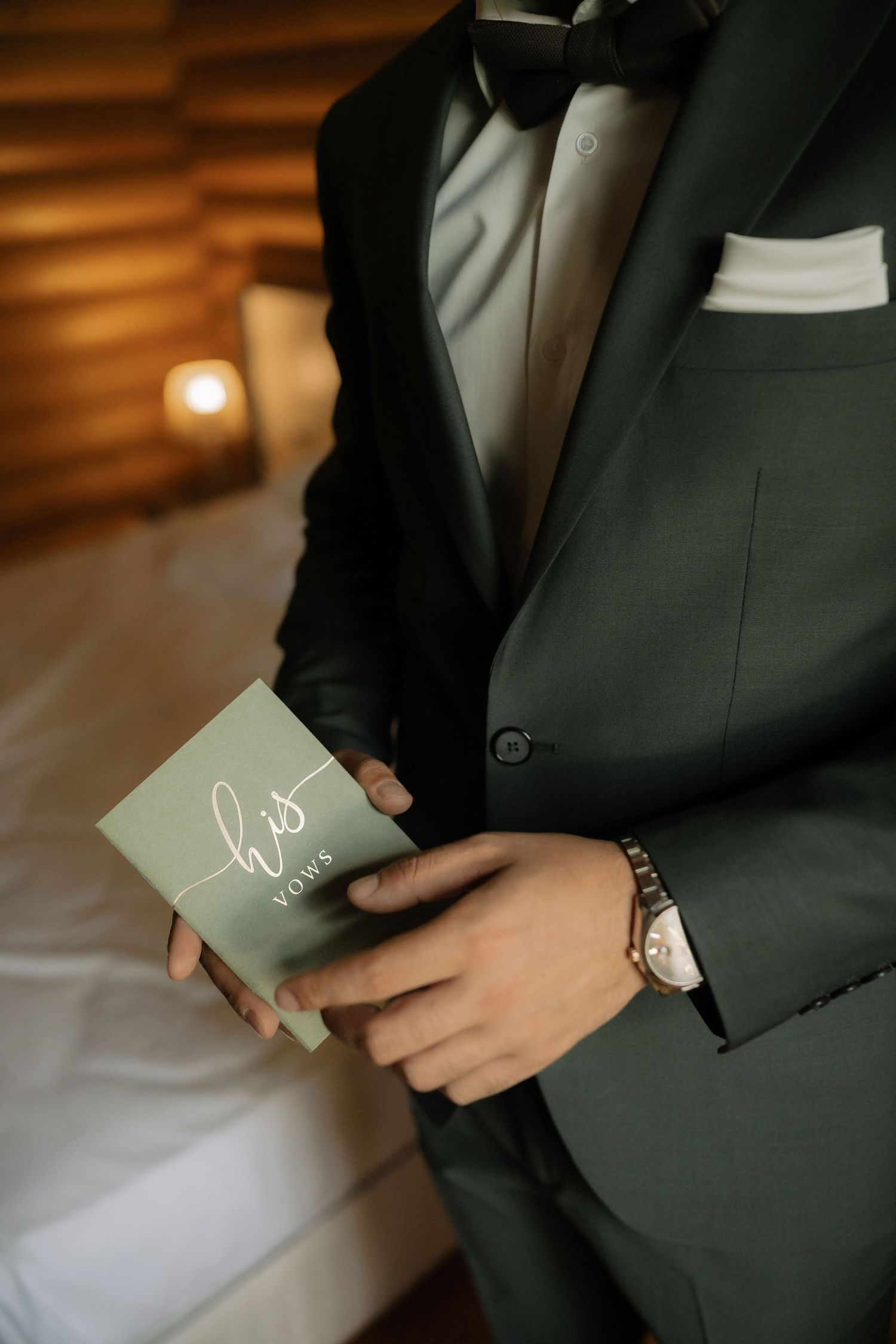 A person in a dark suit holds a small mint green wedding vows book against a blurred warm background.