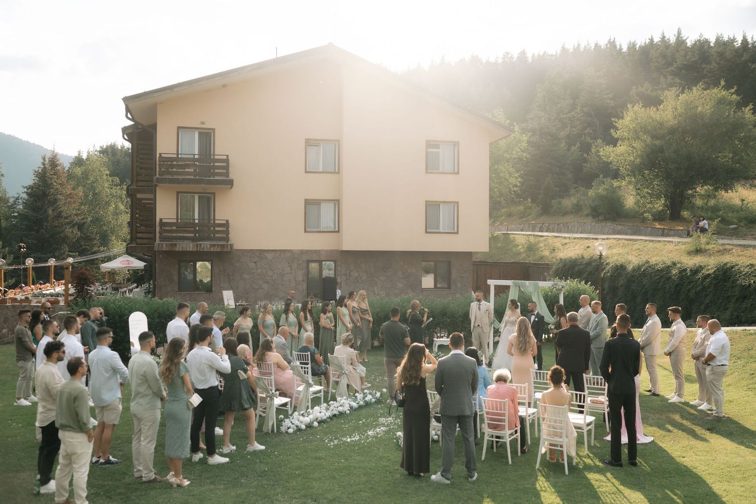 Wedding guests gather outdoors in rows on a grassy lawn with buildings and trees in the background.