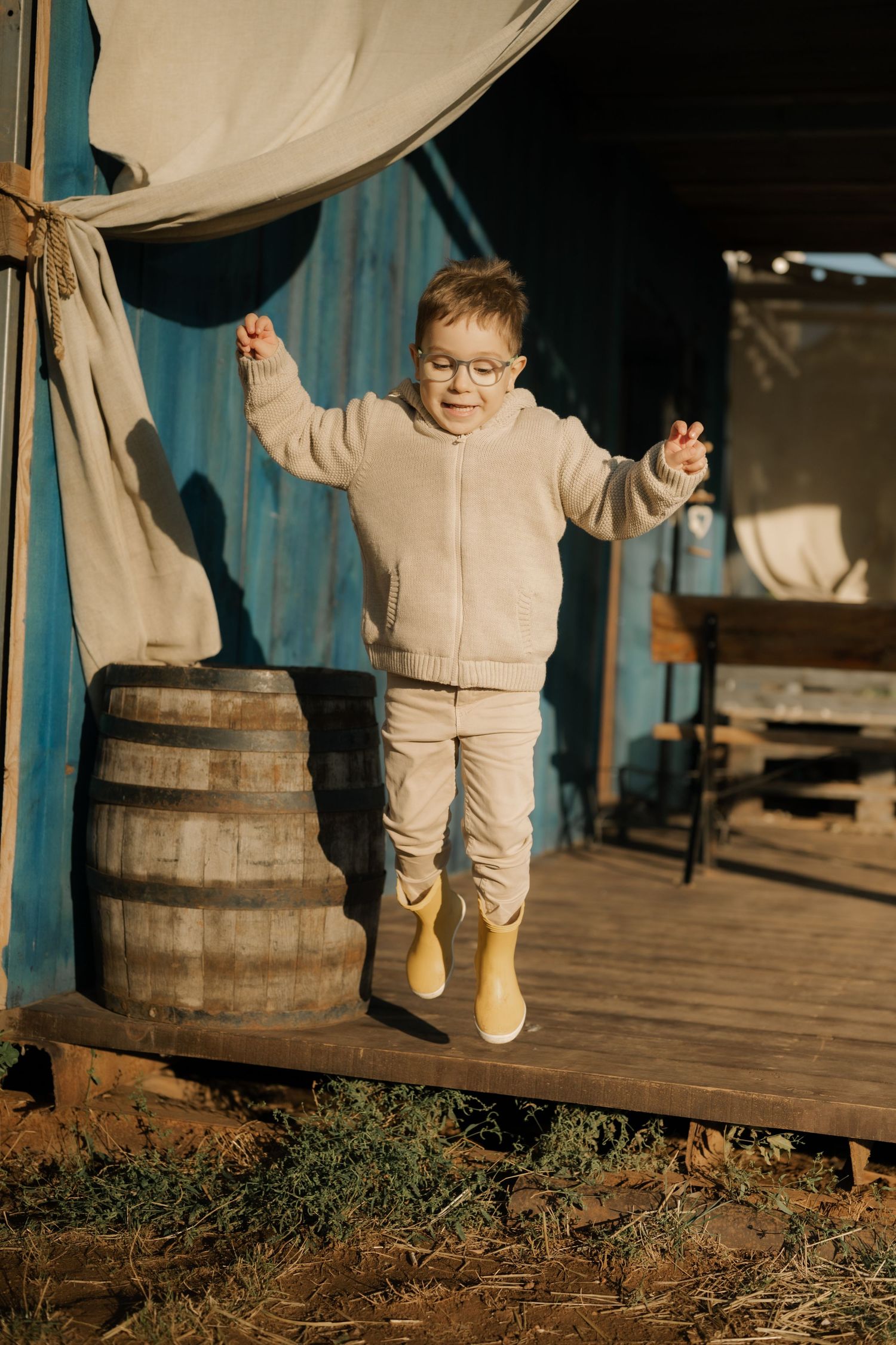 A small child in beige clothing walks along a wooden deck near a rustic barrel in warm sunlight.