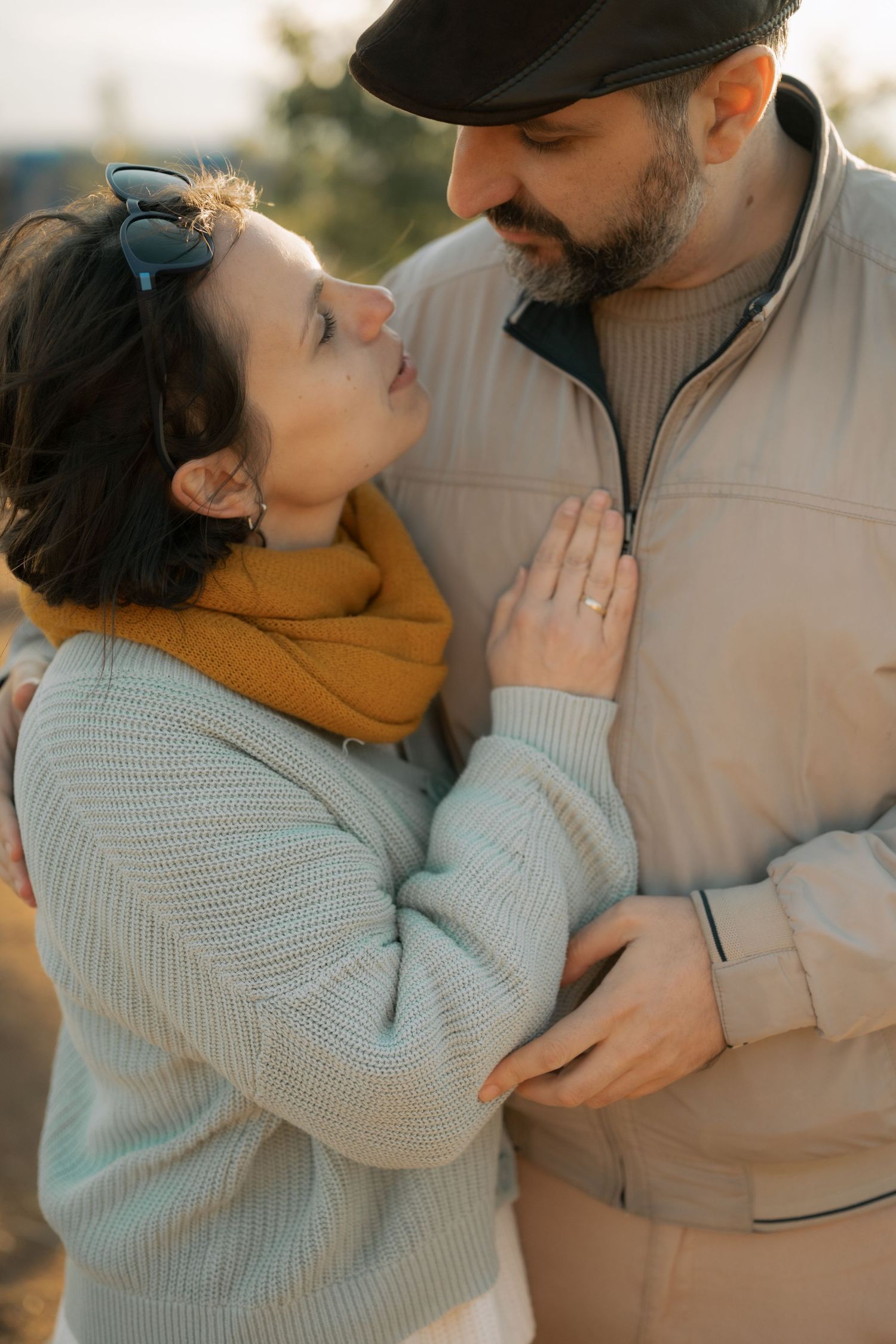 A couple in warm sweaters share an intimate moment outdoors during golden hour, with her hand resting on his chest.