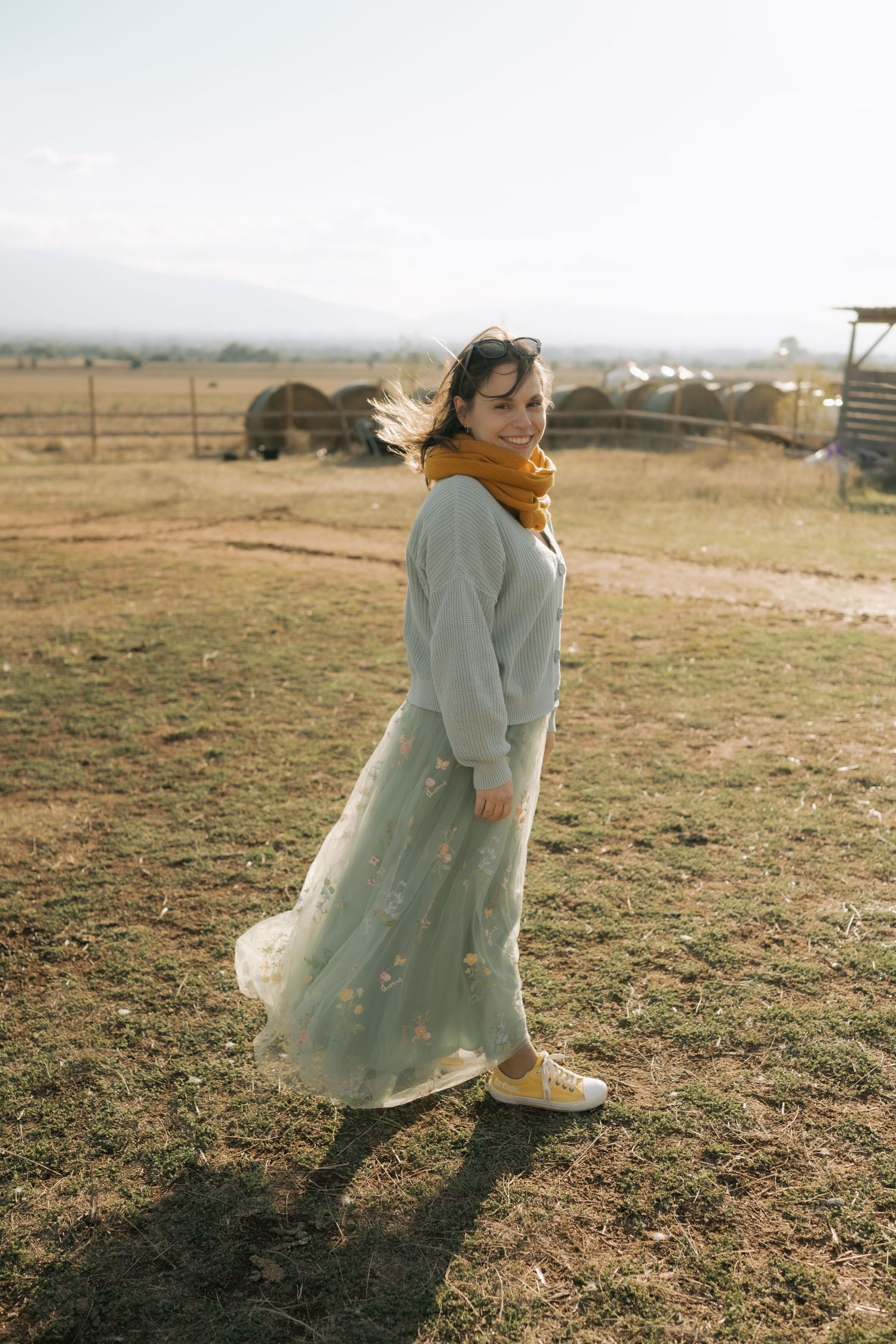 A person in a flowing mint green dress and orange scarf stands in a rural field during golden hour.