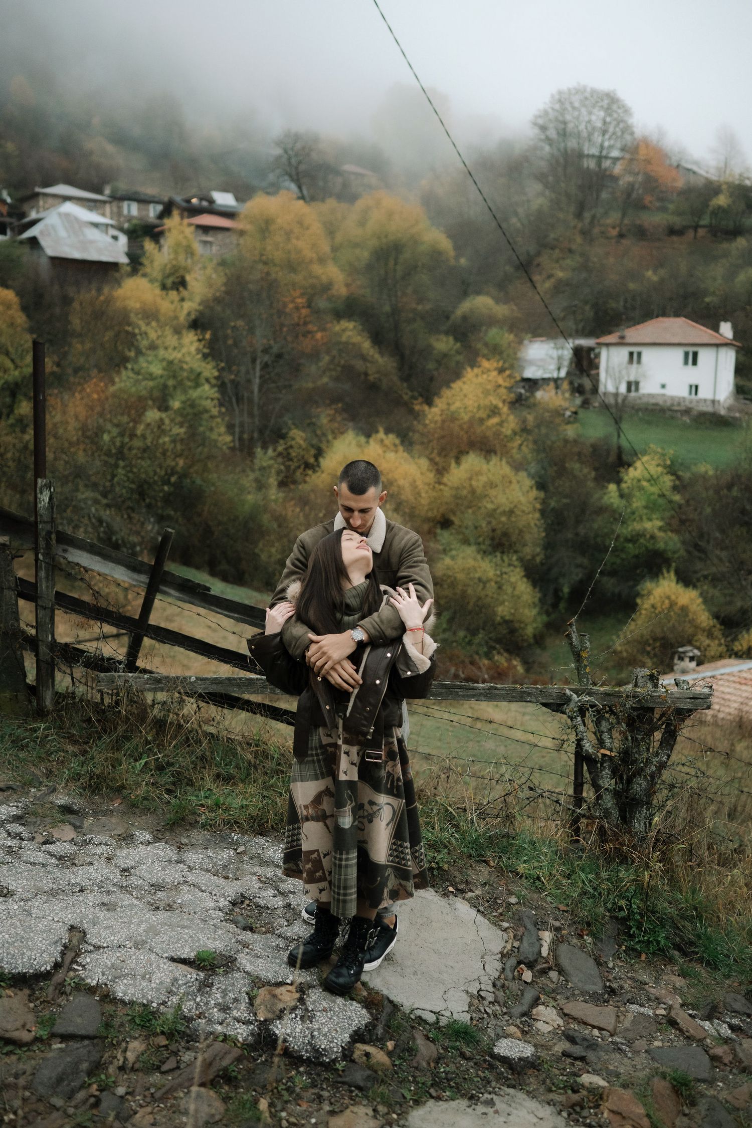 A couple in warm winter coats stands on a misty hillside overlooking autumn trees and houses in a rural setting.