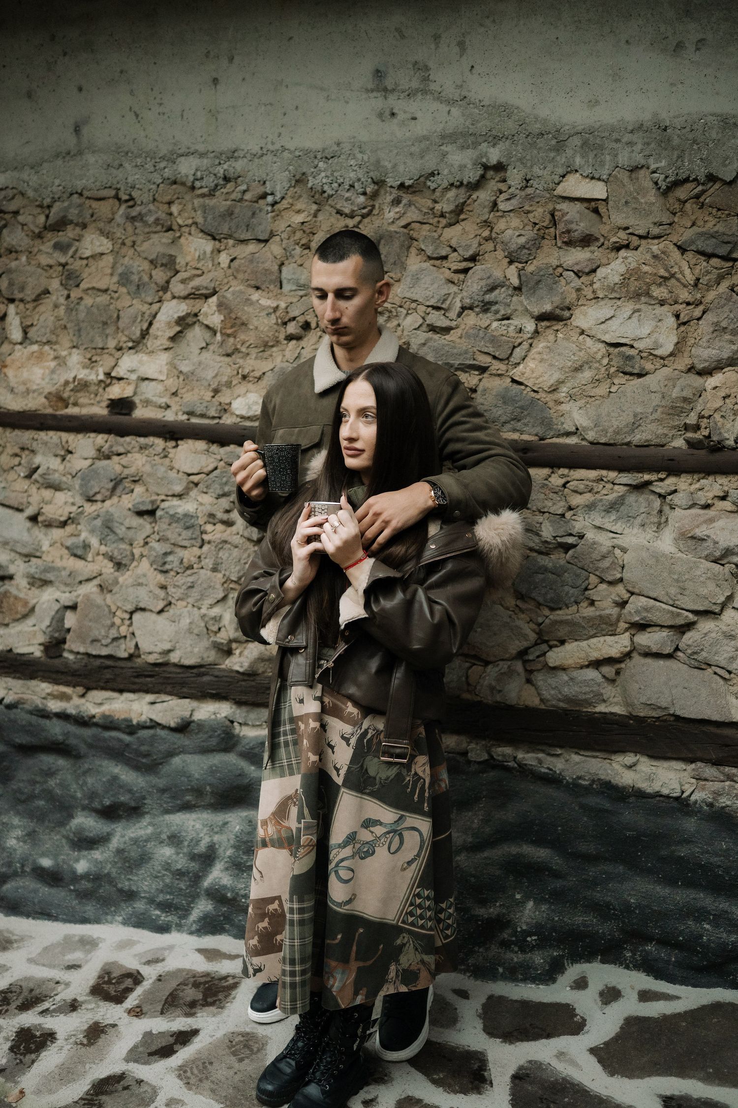A couple poses against a rustic stone wall in a series of moody, atmospheric engagement photos.