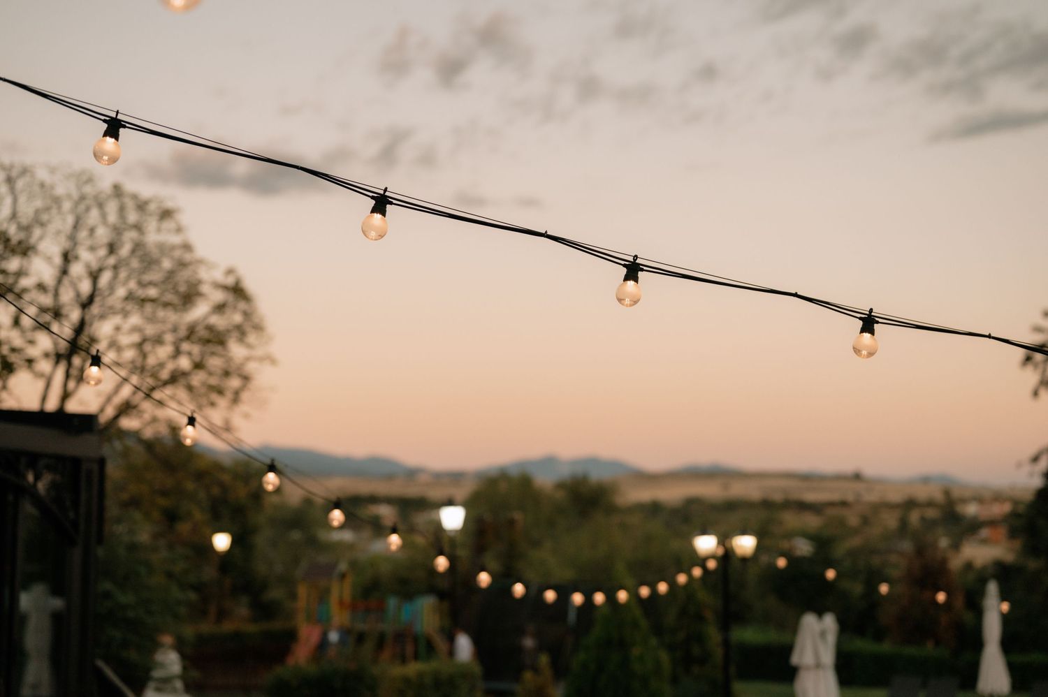 String lights hang against a dusky sky at an outdoor event with mountains visible in the background.