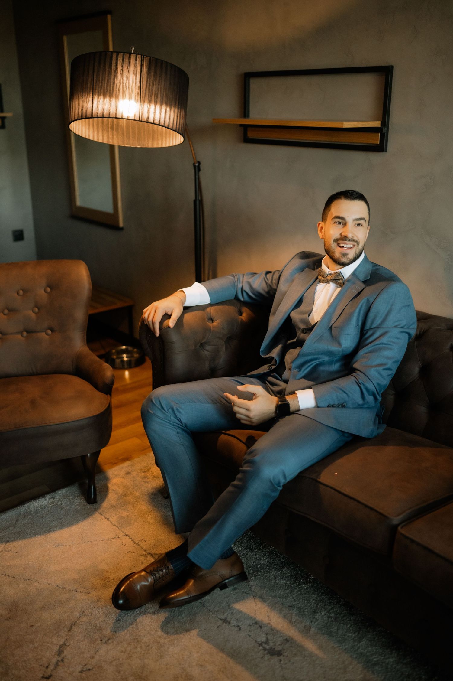 Professional in a navy blue suit relaxes on a brown leather couch while checking their mobile phone in moody lighting.