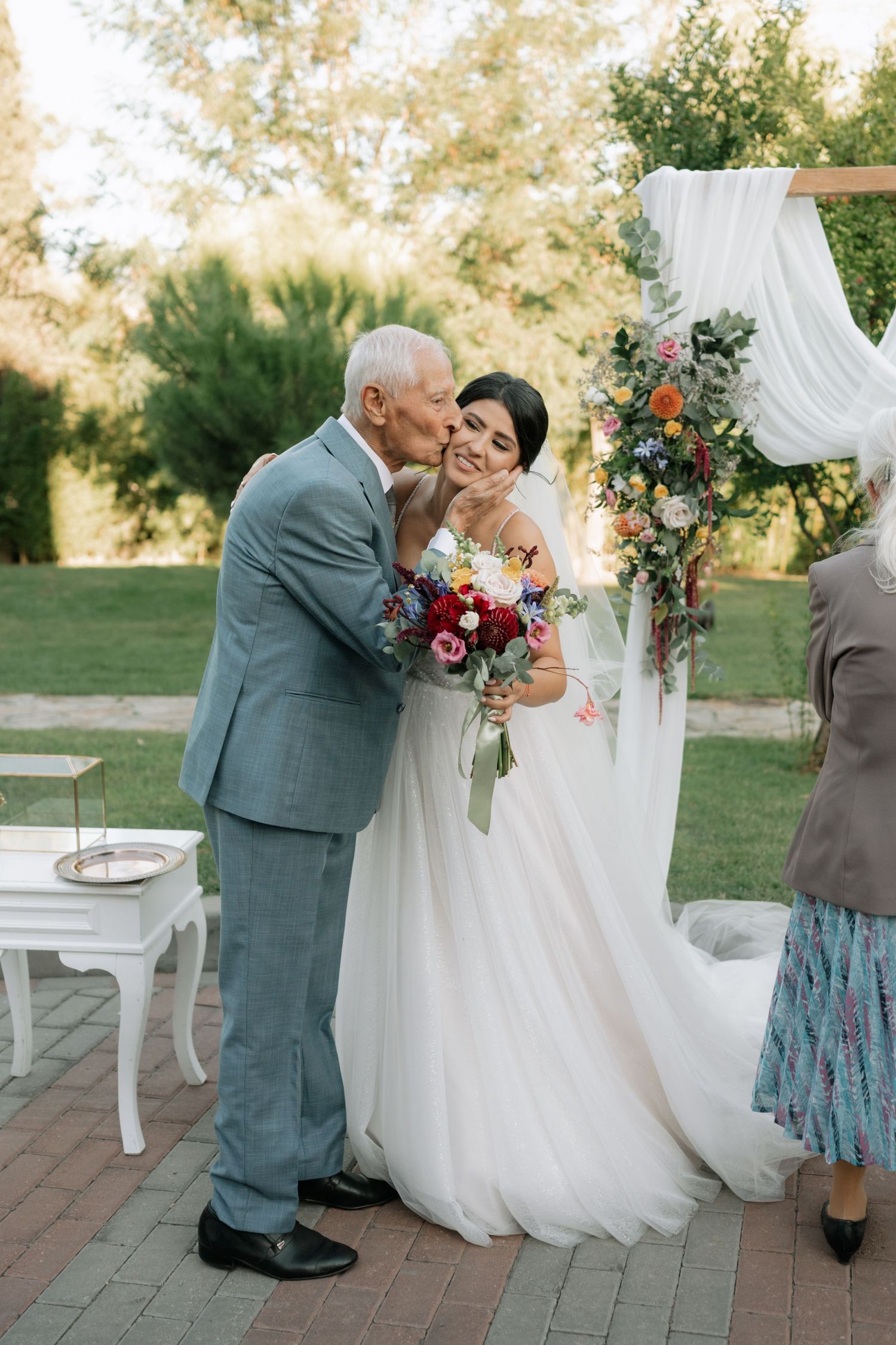 A tender moment at an outdoor wedding ceremony with a white floral arch and decorative drapery on a brick patio.