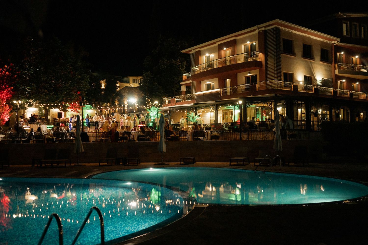Illuminated resort hotel at night with glowing swimming pool and outdoor dining area reflecting in calm water.
