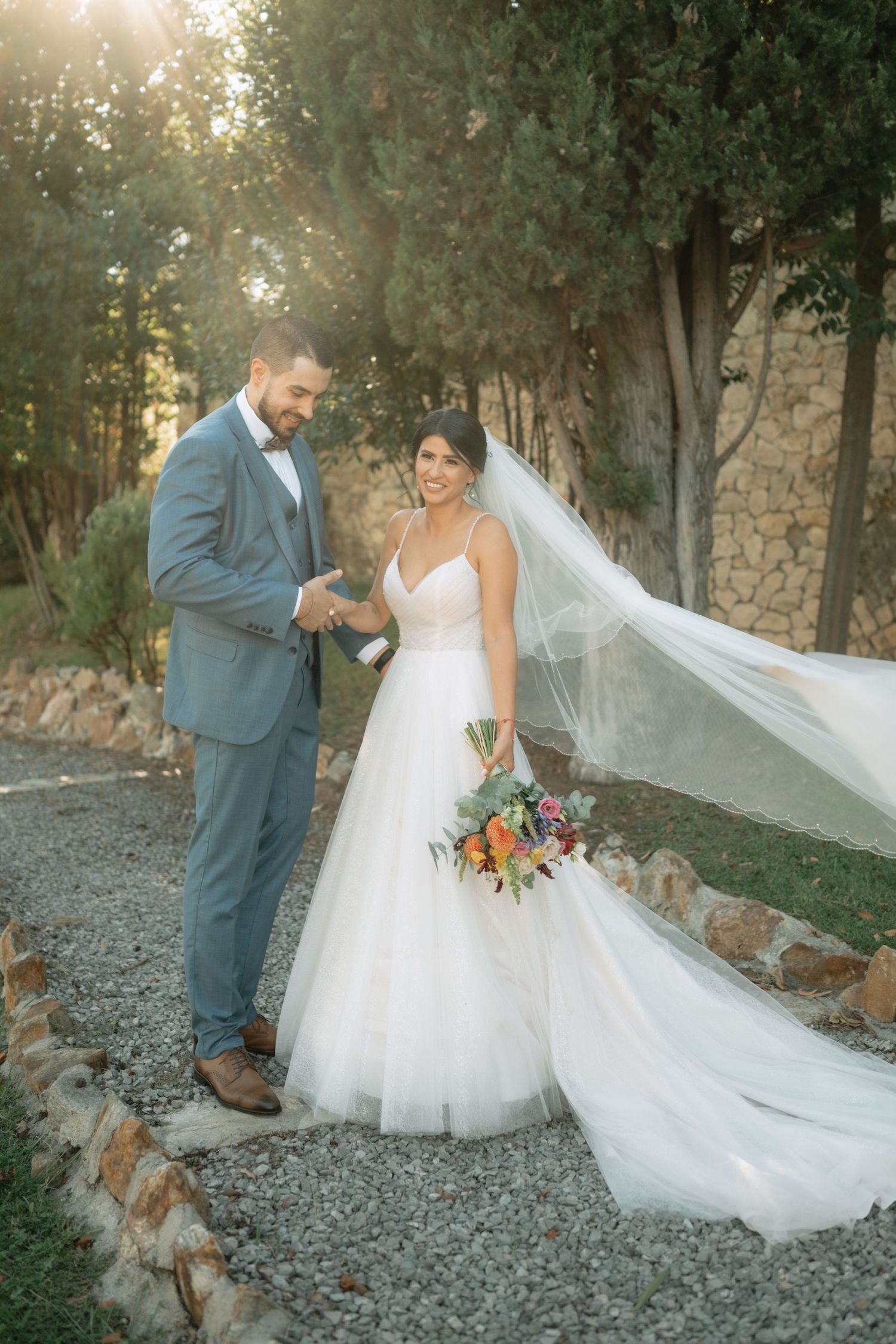 A romantic wedding couple poses outdoors on gravel path with flowing veil and colorful bouquet as their dog joins the moment.