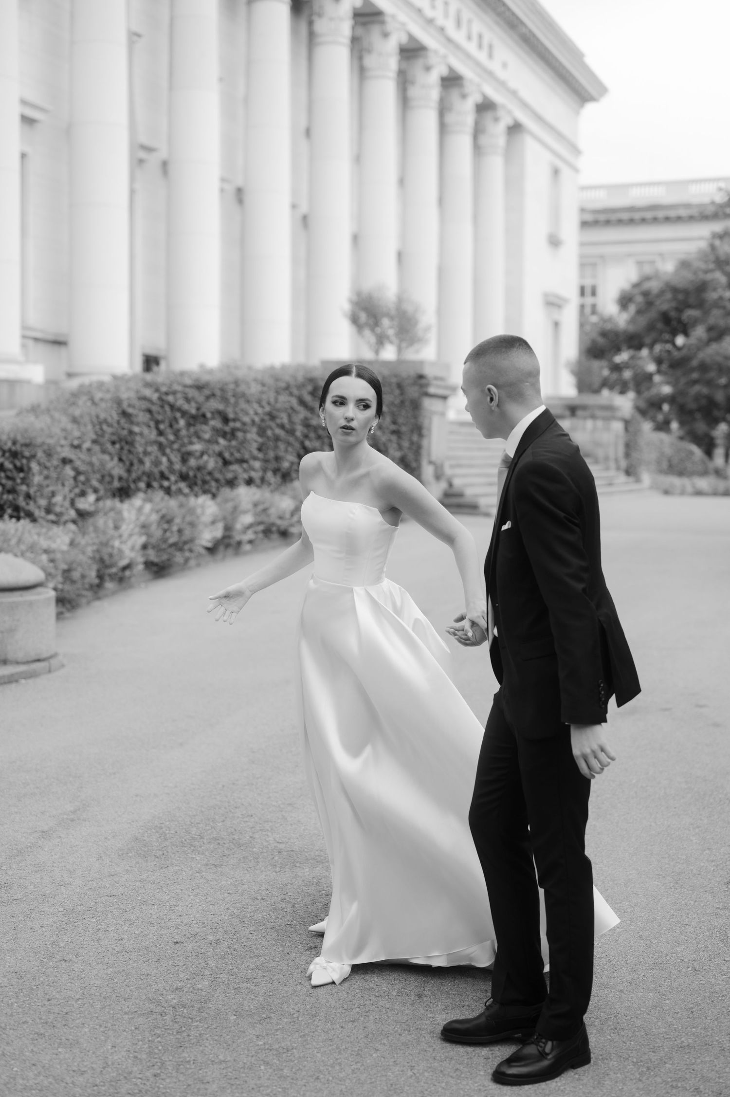 A bride in an elegant white dress twirls as her groom watches outside a grand columned building in black and white.