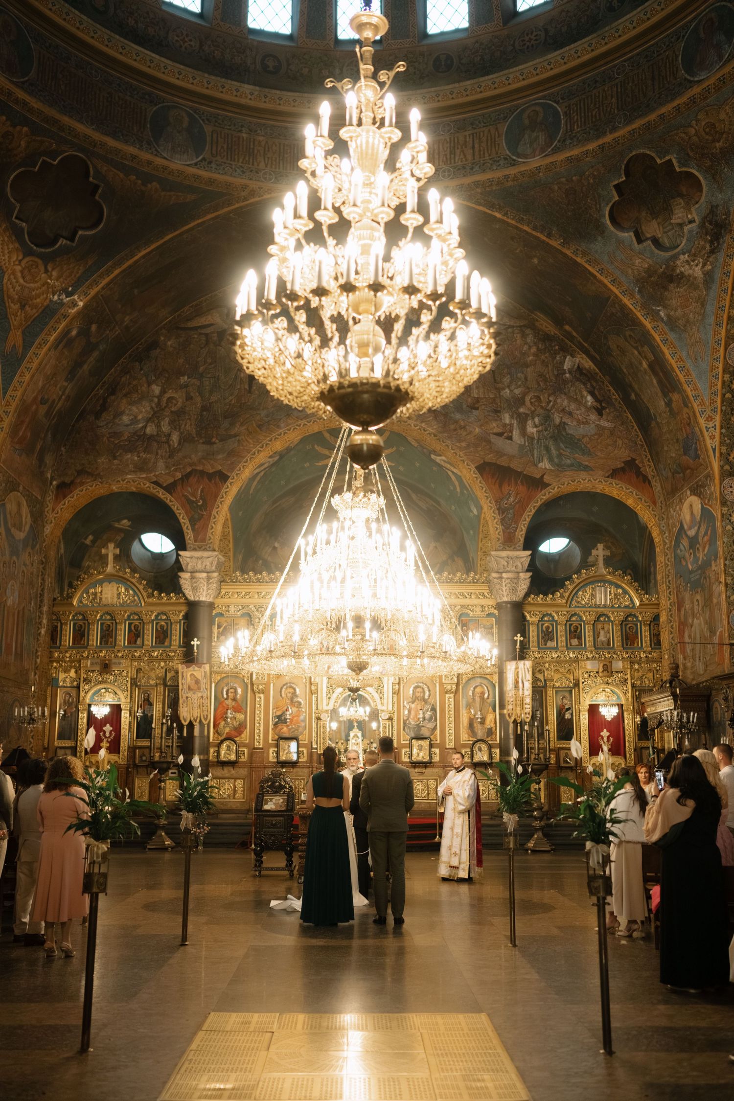 Ornate Orthodox church interior with crystal chandeliers, gilded iconostasis, and decorated dome ceiling during a service.