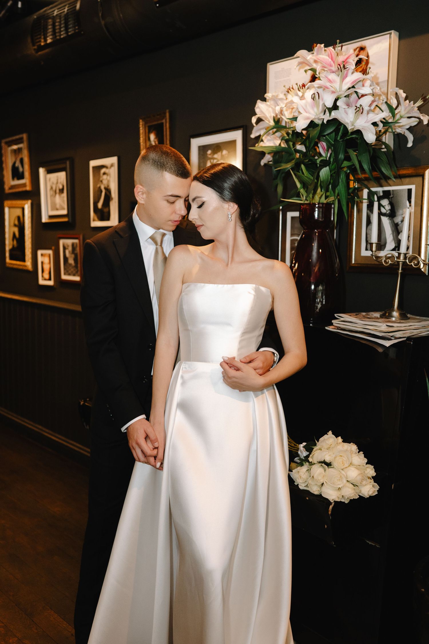 A romantic wedding moment captured in an art gallery with dark walls and framed portraits in the background.