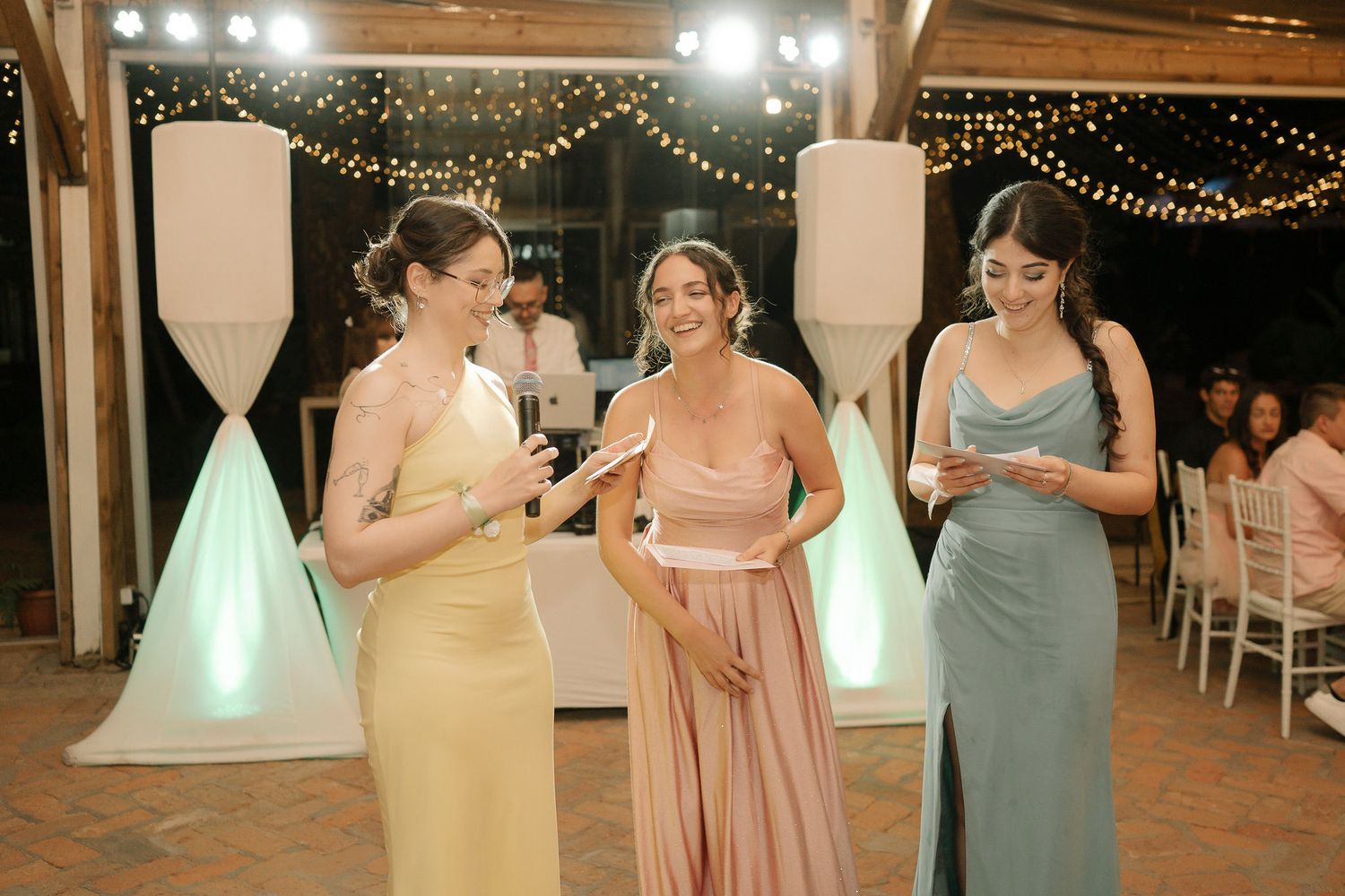 Bridesmaids in pastel pink and gray dresses dance together during a wedding reception under string lights.