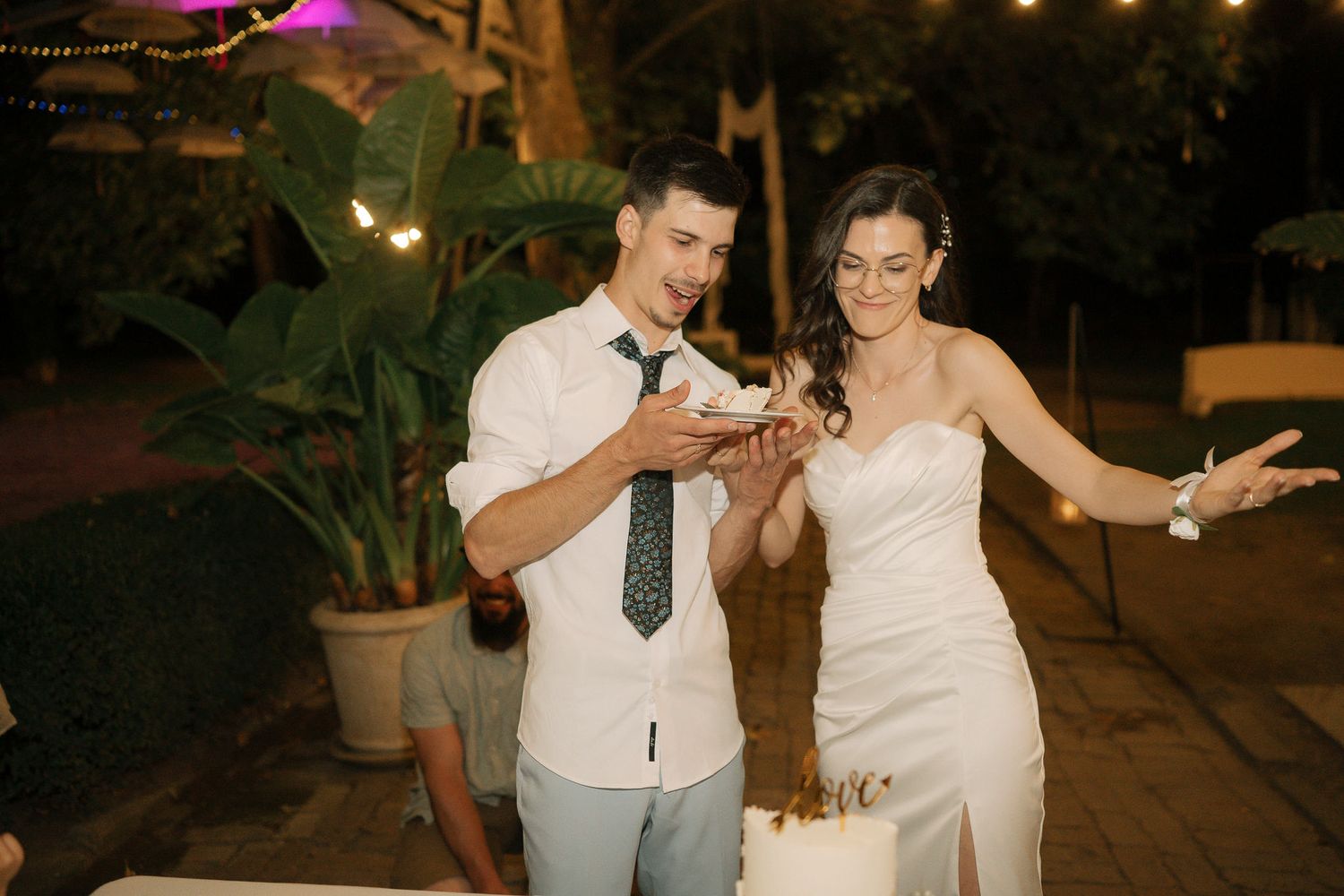 A couple shares a romantic moment while cutting their wedding cake at an outdoor evening reception.