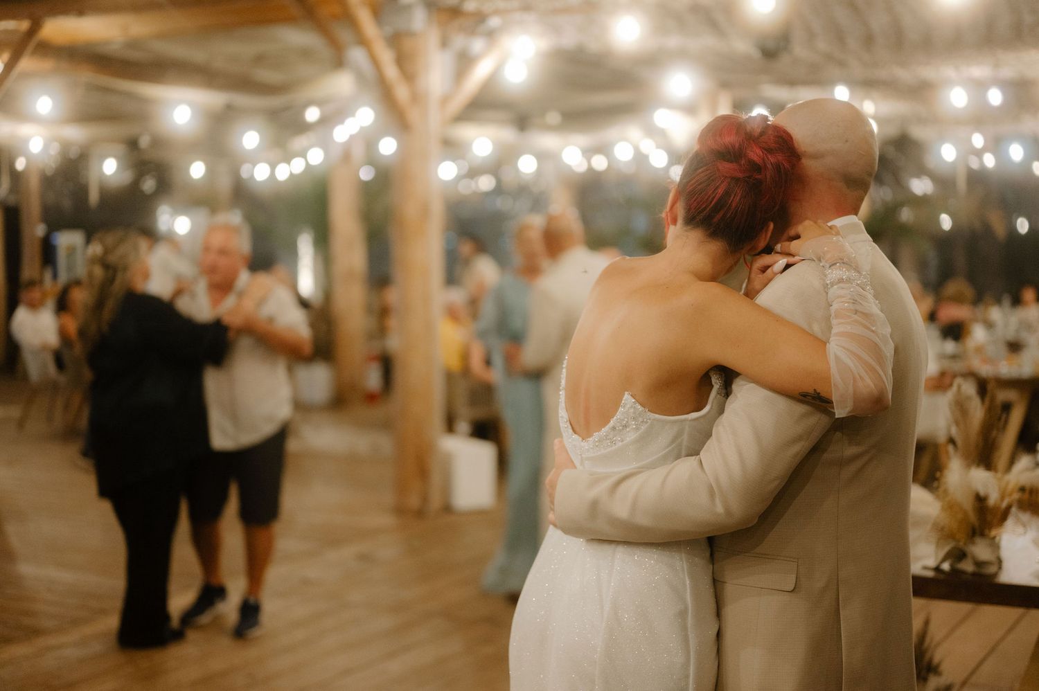 Couples slow dance at a rustic indoor wedding reception venue decorated with string lights and wooden beams.