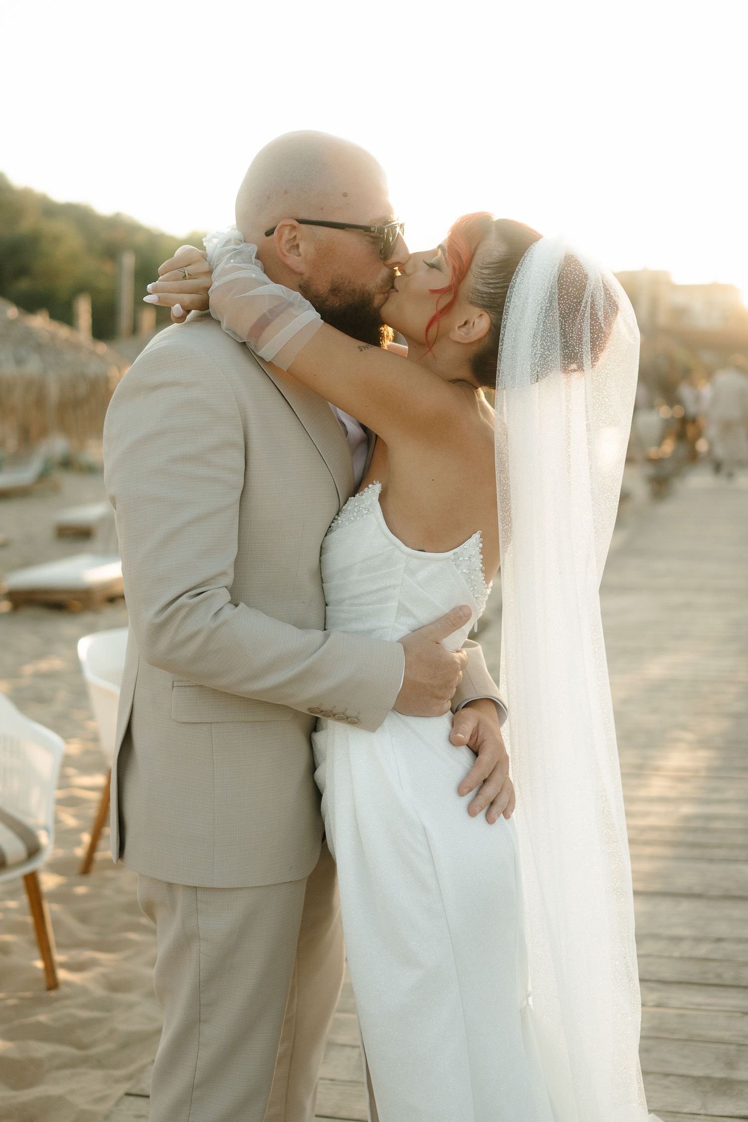 Newlywed couple shares romantic kiss at sunset on beach boardwalk with boats in background.