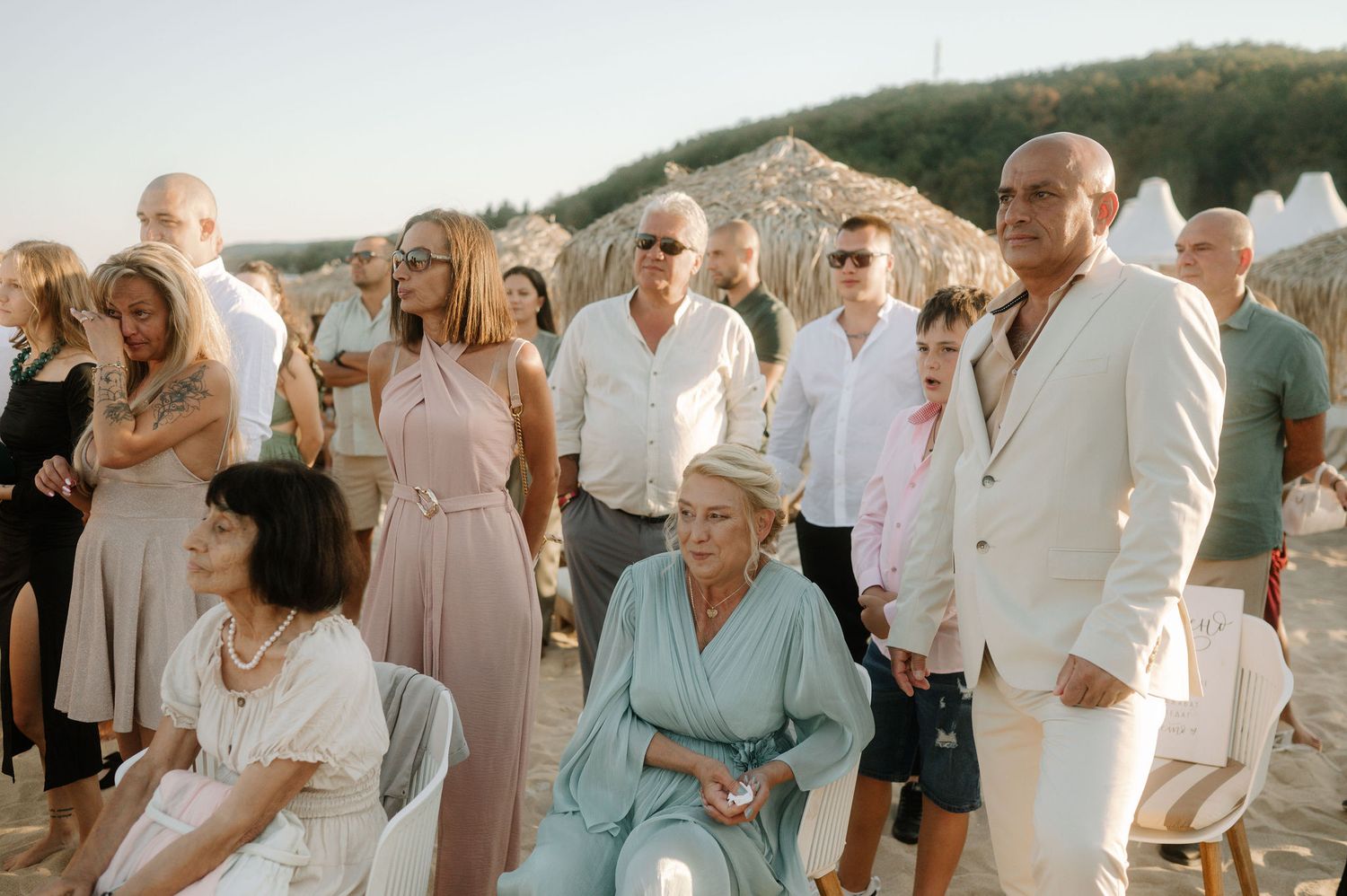 A group of wedding guests in light summer attire gather outdoors during a sunset celebration.