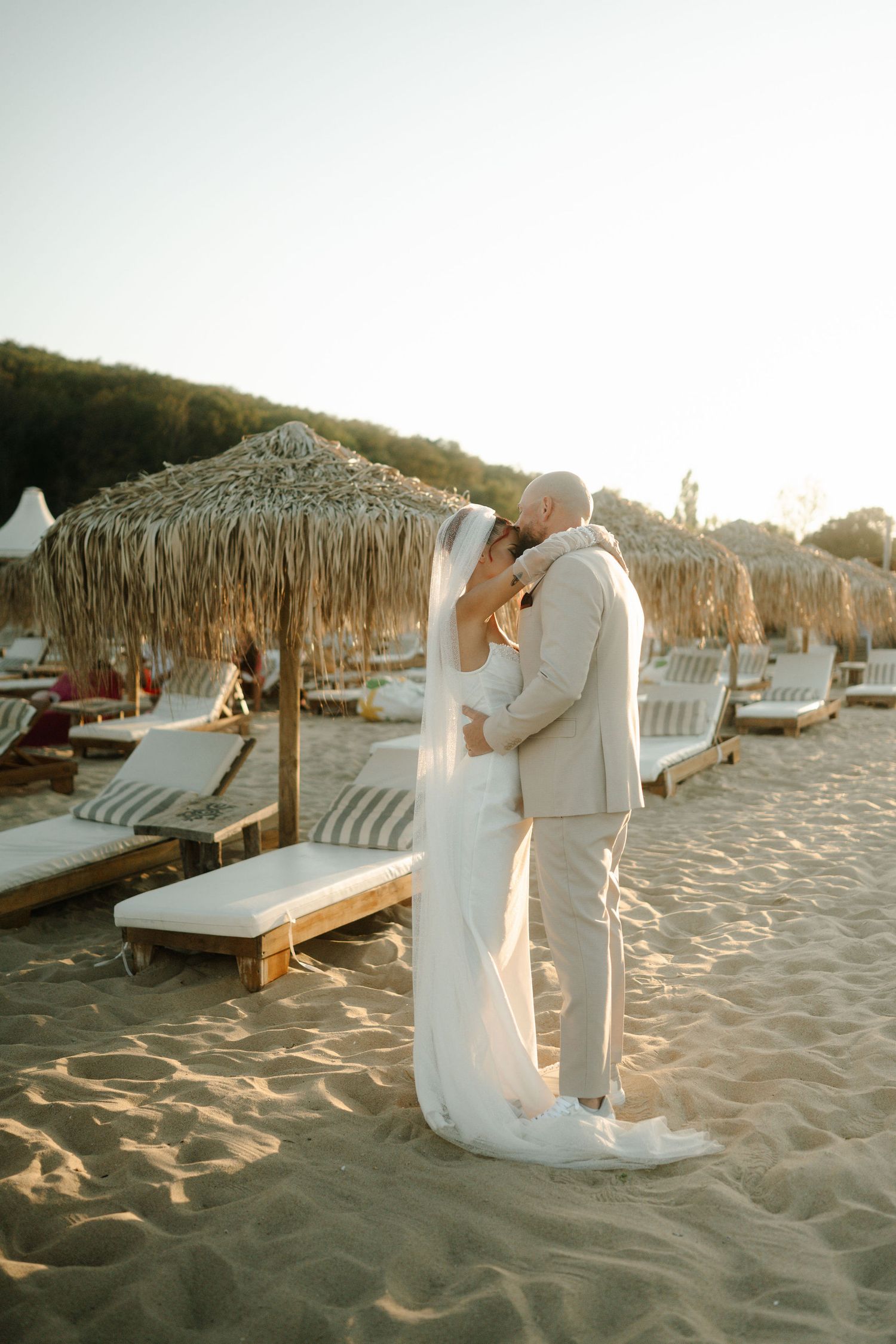 A romantic beach wedding scene at sunset with thatched umbrellas and lounge chairs on the sandy shore.