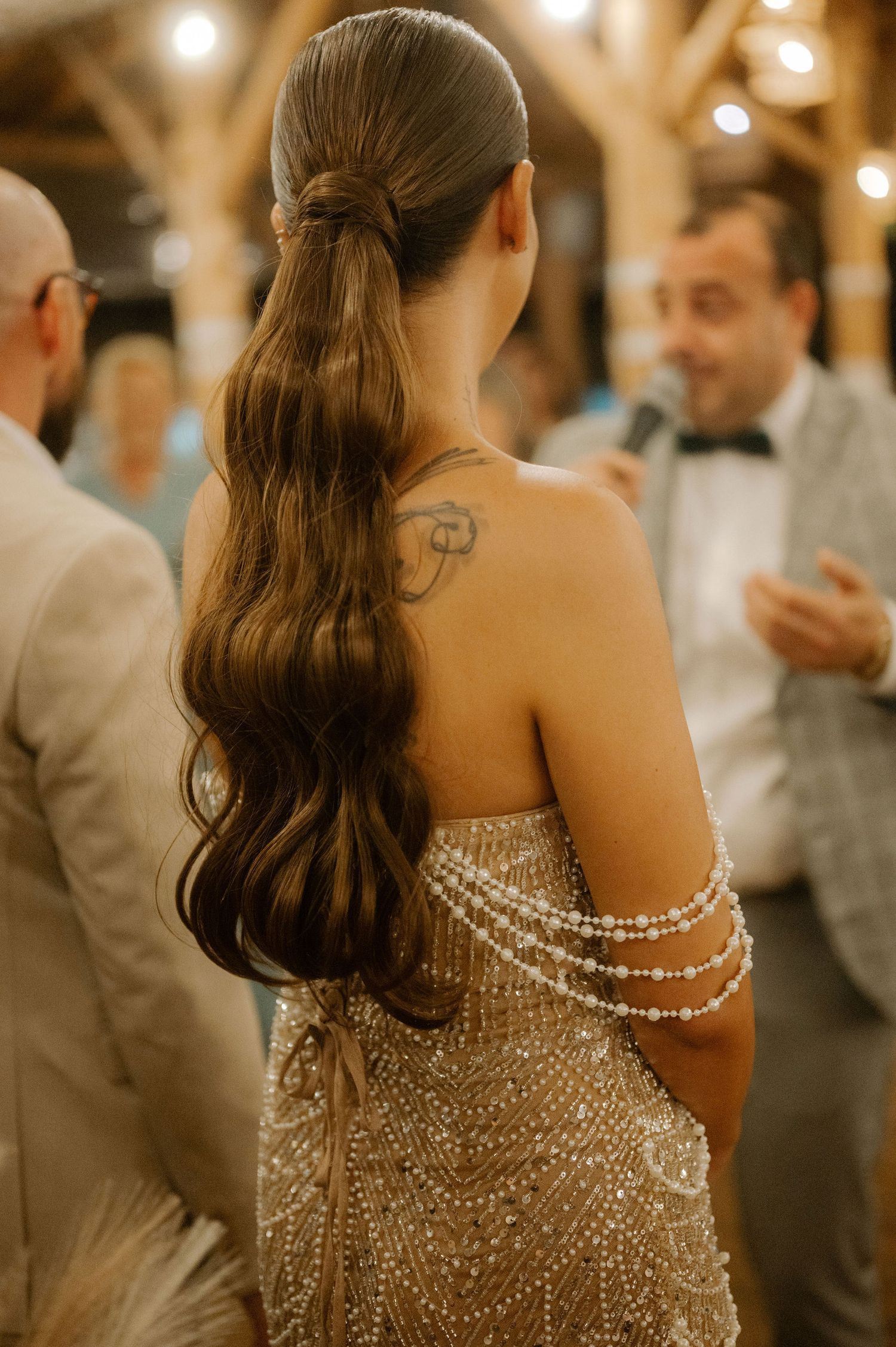 Back view of a bride in a sparkly off-shoulder gown with long wavy hair in a ponytail at an elegant reception.