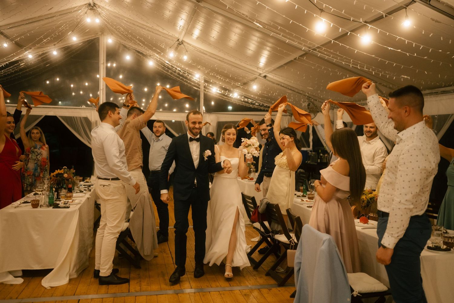 Wedding guests dance and celebrate under warm string lights at an indoor reception venue.