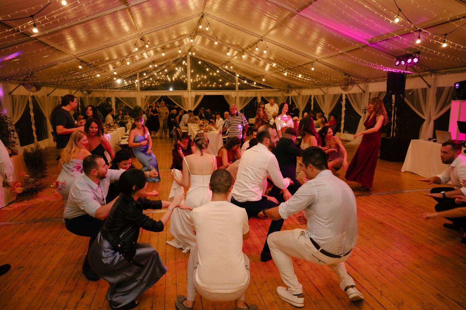 Wedding guests gathered in a circle on a dance floor under string lights in a tent venue.