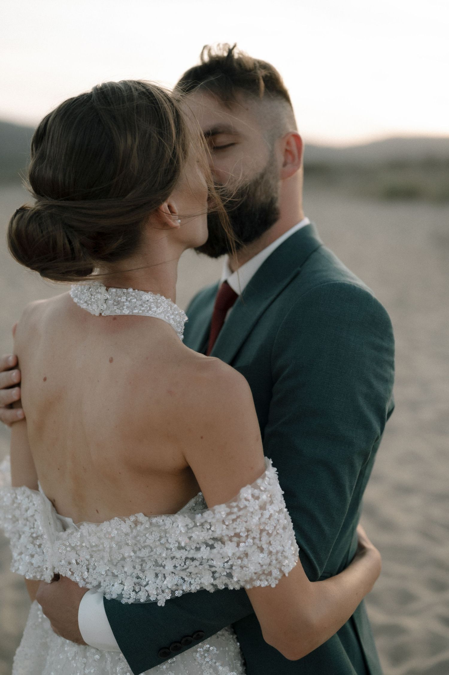 A bride and groom share a tender kiss on a sandy beach at sunset, she in a lace gown, he in a green suit.