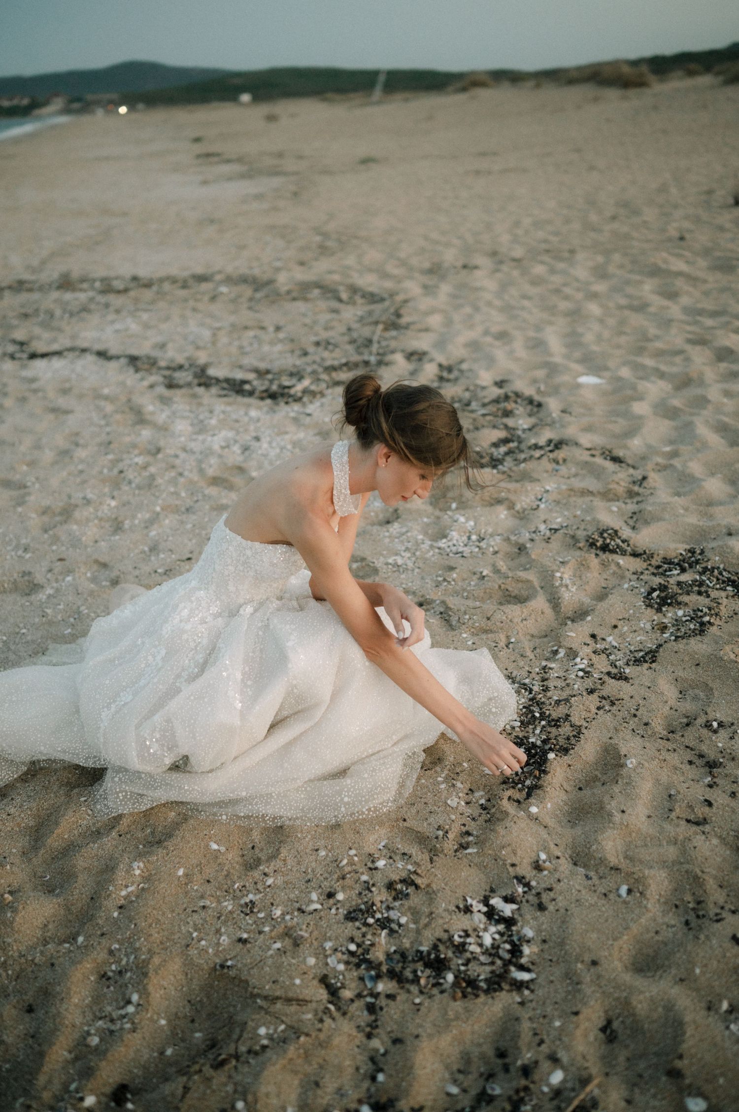 A bride in a white gown sits on sandy beach at dusk, gently touching scattered shells and pebbles.