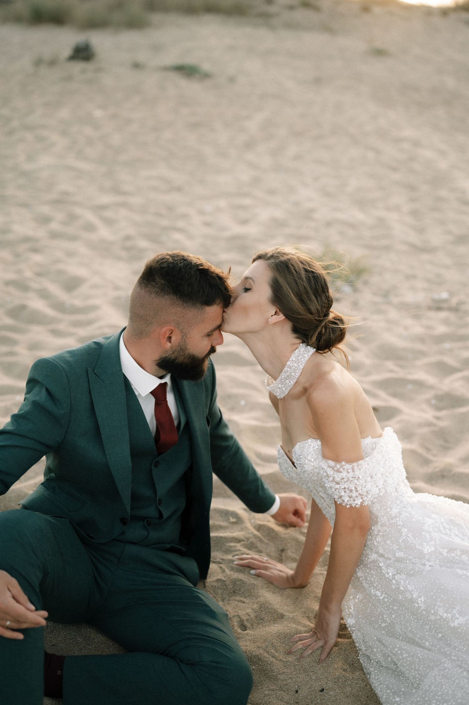 Bride in white lace dress kissing groom in green suit on sandy beach during romantic outdoor wedding photo shoot.