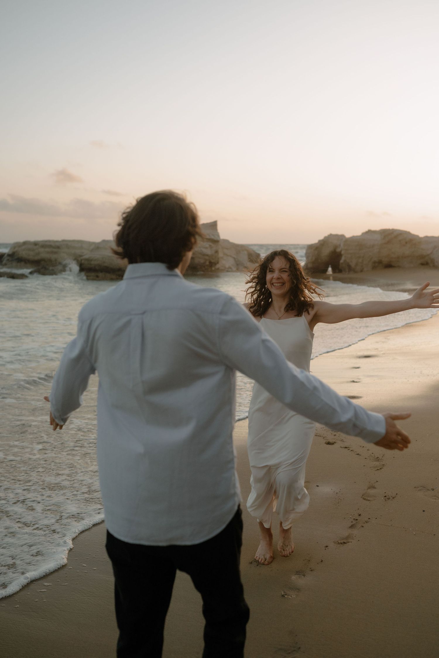 Couple running toward each other on a sandy beach at sunset, woman in white dress, man in white shirt, ocean waves behind.